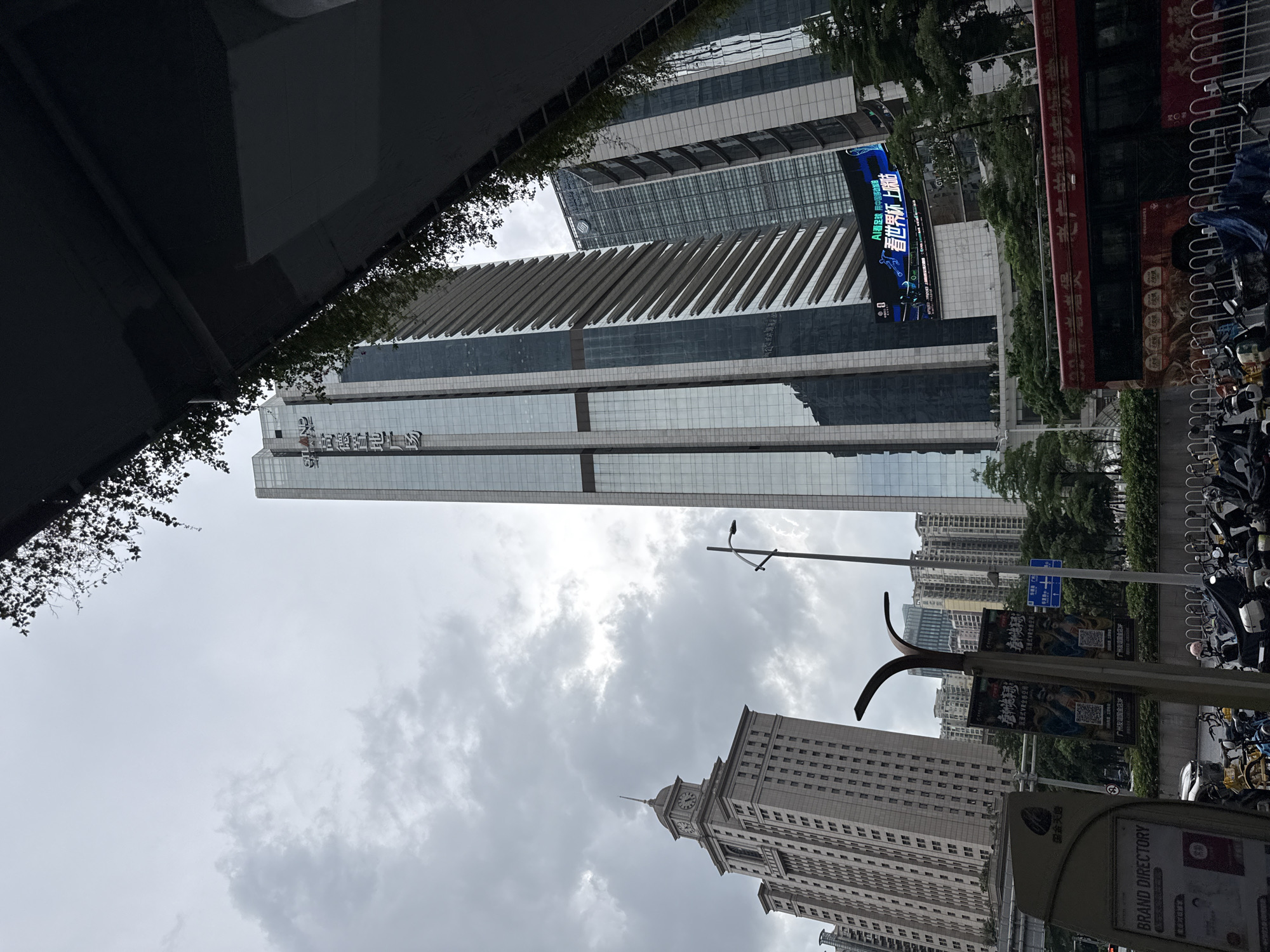 The image captures a street-level view of the Tianhe District in Guangzhou, China, under an overcast sky. Dominating the skyline are several modern skyscrapers with glass facades, including the tall, slender building with a distinctive stepped design on its right side. To the left, a lighter-colored building with a clock tower is visible. In the foreground, a red bus with Chinese text and imagery is parked behind a white metal fence. Several motorcycles and scooters are parked along the sidewalk. A large digital billboard displays a promotional advertisement, featuring Chinese text and graphics. The scene suggests an urban environment with active commercial and transportation activity.