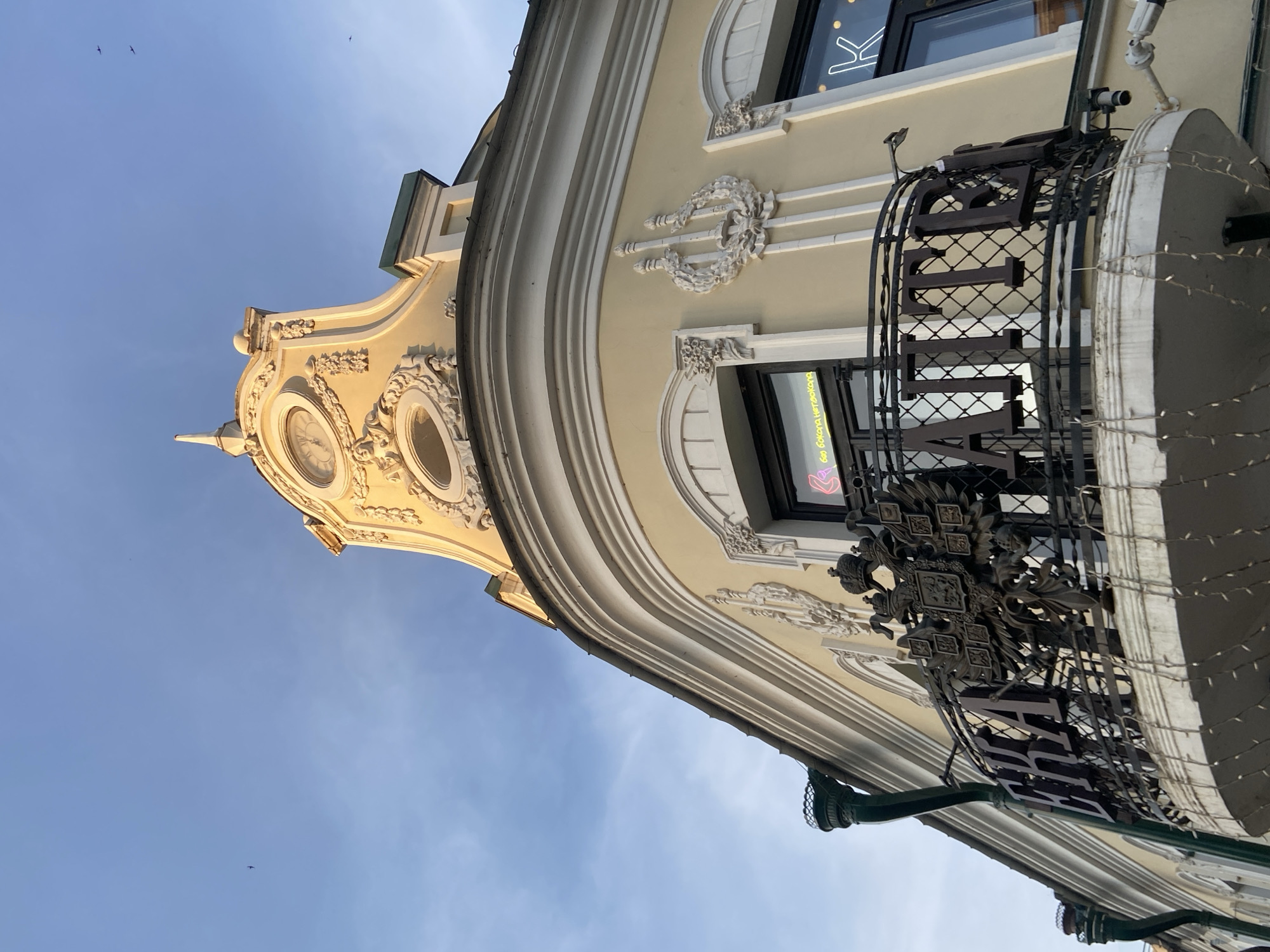 A low-angle, upward-tilted shot features a section of ornate architecture in Kazan, Russia. The building's facade, finished in a pale yellow hue, showcases neoclassical ornamentation. The architectural details include curved sections, decorative wreaths, and circular windows. A black, wrought-iron balcony with the Cyrillic word "ВИНТАЖ" (Vintage) is present. The blue sky is visible in the background, and there are a few small birds flying.