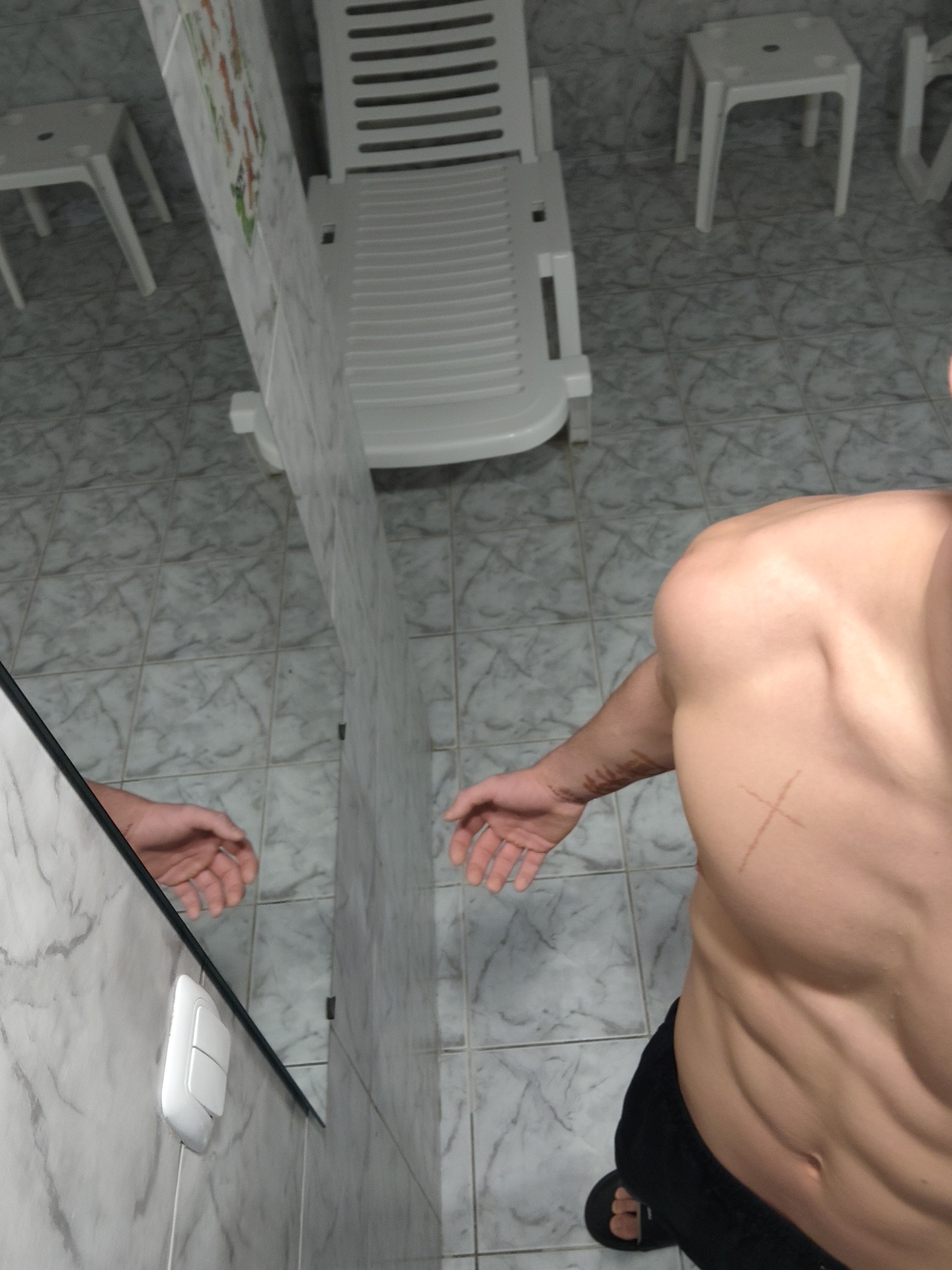 A medium-high-angle shot depicts a person in a room, likely a bathroom, possibly located in Kozloduy, Bulgaria, based on contextual information not provided in the image. The person is partially visible, primarily showing their torso, which is bare-chested, revealing abdominal muscles. They are wearing dark bottoms. A faint X-shaped mark is visible on their upper chest/shoulder area. The person's arm is extended towards the left of the frame, and their hand is partially visible. A reflection in a mirror shows a portion of the person's hand and arm. The background shows white tiled walls, and a white plastic deck chair. Two small white plastic stools are visible in the background as well, to the right and slightly behind the deck chair. The floor is covered in light-colored, patterned tile. A white light switch is visible on the wall near the mirror.