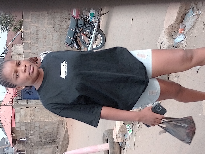 A woman wearing a black shirt and denim shorts stands outdoors. She holds a phone and a black plastic bag in her hand. Behind her is a parked motorcycle and a brick building. The ground appears to be dirt or unpaved. The image is slightly rotated, causing a skewed perspective.