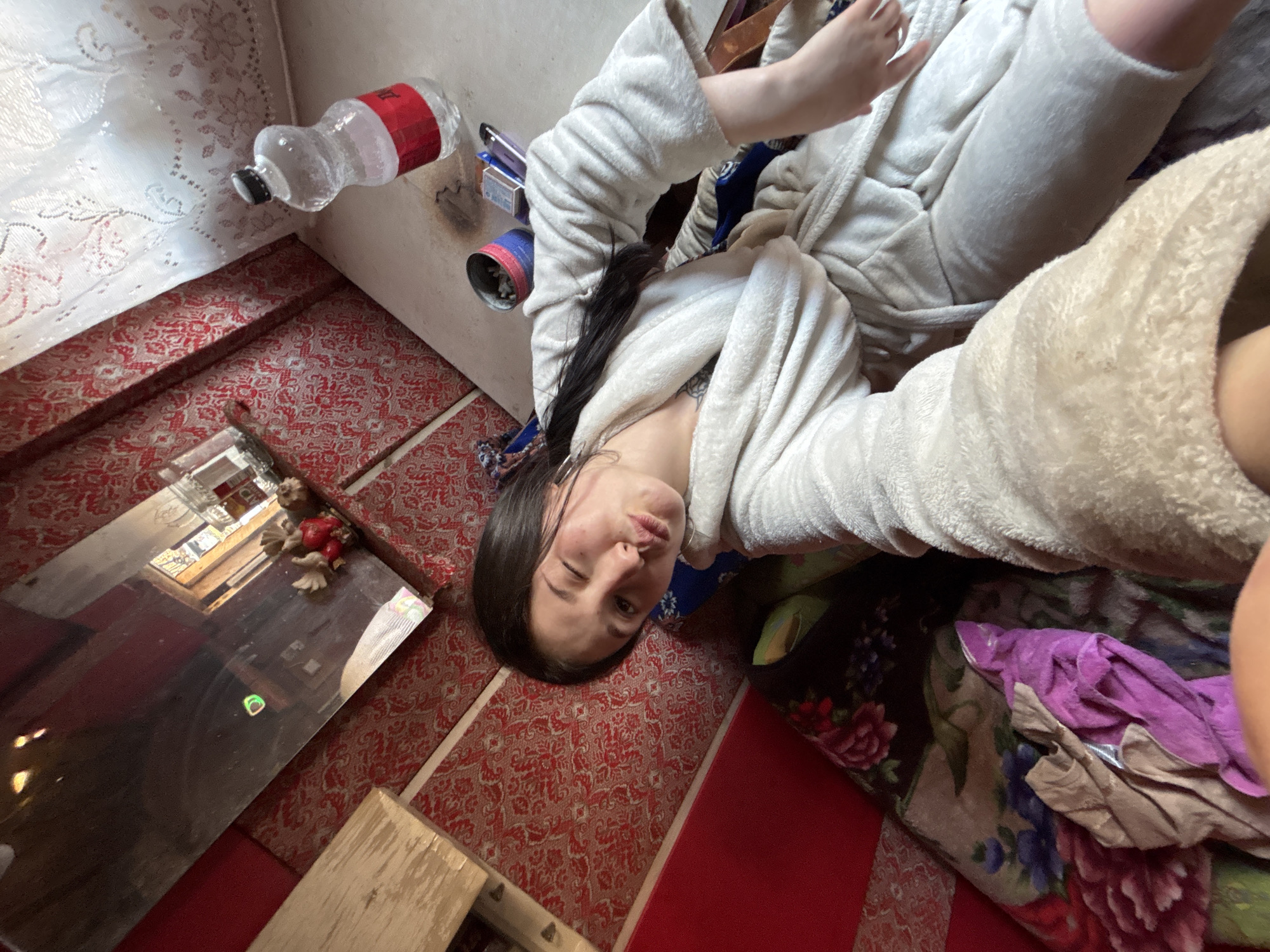 The image, taken in the city of Фоки, Russia, appears to be a selfie of a young woman in a white robe. She is partially visible, positioned diagonally across the frame, winking at the camera. Her dark hair is pulled back.
The setting is a cluttered room with red patterned wallpaper and carpeting. In the upper left corner, a plastic water bottle and various containers sit near the wall. A mirror is on the floor, reflecting a window and items within the room. Other visible objects include miscellaneous items on the bed and a wooden structure in the lower left.