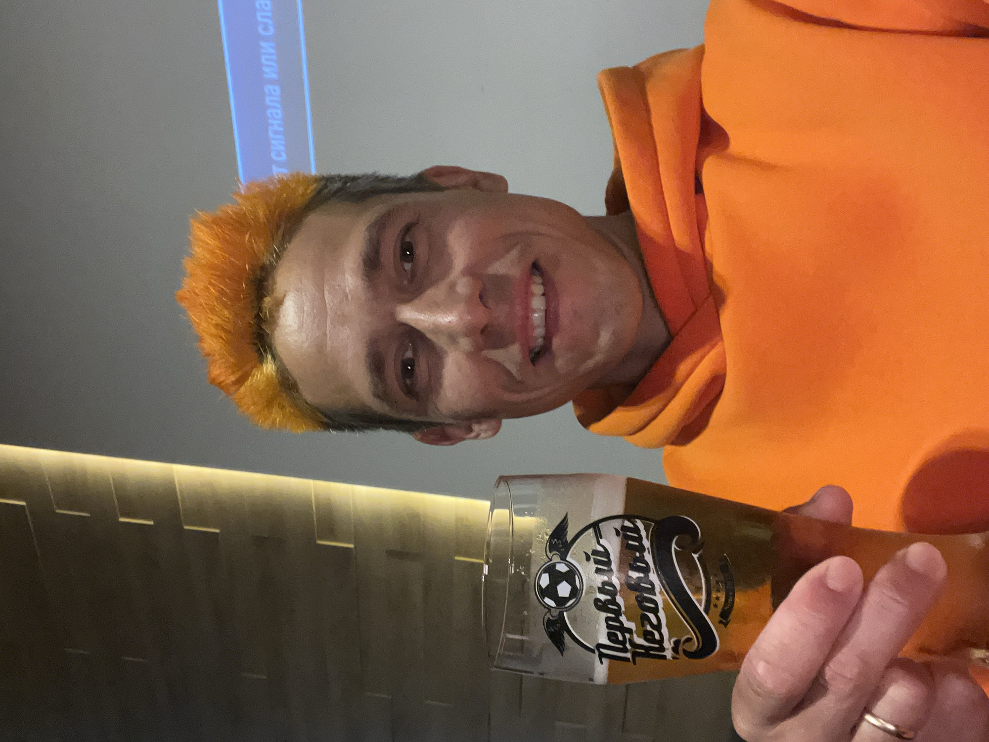 An image, rotated ninety degrees clockwise, shows a man smiling and holding a glass of beer in an indoor setting. The man, appearing to be in his 30s or 40s, has short, distinct bright orange hair on top, with darker hair on the sides. He is wearing a vibrant orange hooded sweatshirt and is looking directly at the camera with a broad, cheerful smile, showing his teeth.
He holds a tall glass of amber-colored beer, topped with a small amount of foam, in his right hand. The glass features a decorative logo that includes a stylized football with wings, and the Russian text "Первый Незовый" (Pervyy Nezovyy). A wedding band is visible on his left ring finger.
The background is dimly lit. On the upper left, a light-colored surface, possibly a screen or wall, displays partially visible blue Russian text, reading "Т сигнала или сла" (likely part of "Нет сигнала или слабый," meaning "No signal or weak"). On the lower right, a darker wall features a horizontal strip of warm, indirect lighting. The overall scene suggests a casual evening at a bar or similar establishment, likely in Chaikovsky, Russia, given the context and visible text. The man's expression conveys happiness and enjoyment.