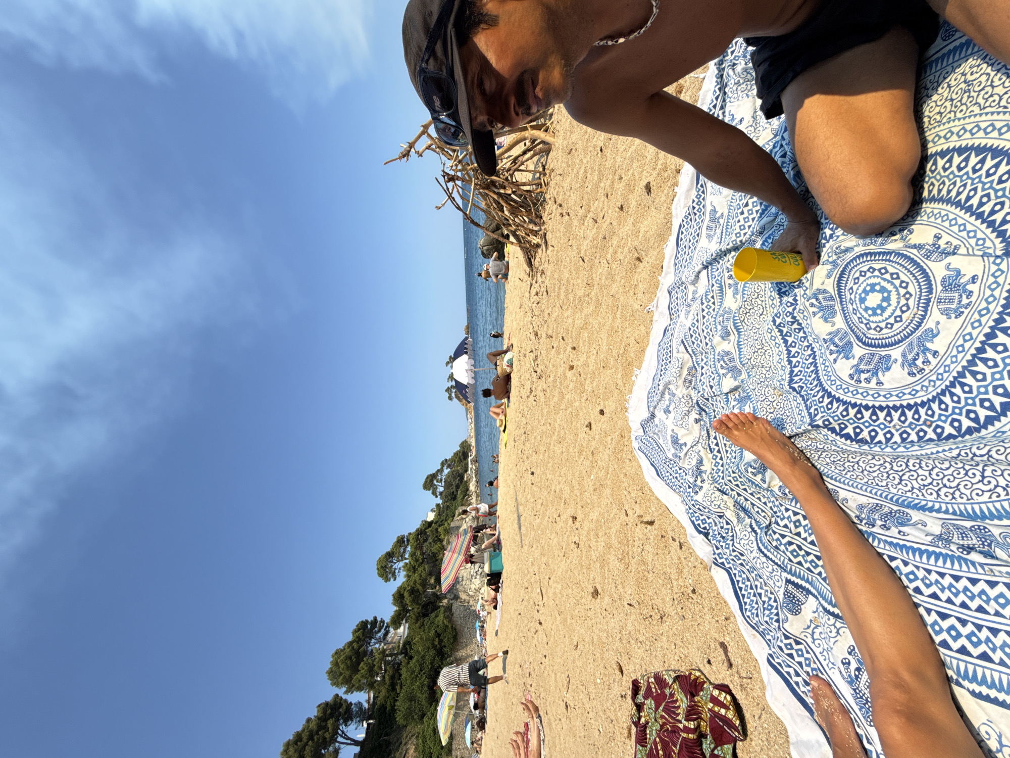 A bright, sunny day at a crowded beach in Calonge i Sant Antoni, Spain. The foreground features a man in a black hat and sunglasses relaxing on a blue and white patterned beach towel, holding a yellow cup. To his left, a person's legs are visible, also on the towel. The sandy beach is populated with many people sunbathing, some under umbrellas. In the background, the Mediterranean Sea is visible, with a large, white cruise ship partially submerged or listing on its side near the shore. Lush green trees and buildings are situated on a hillside overlooking the beach. A haphazard structure made of driftwood is also present on the sand. The sky is a clear blue with scattered white clouds.