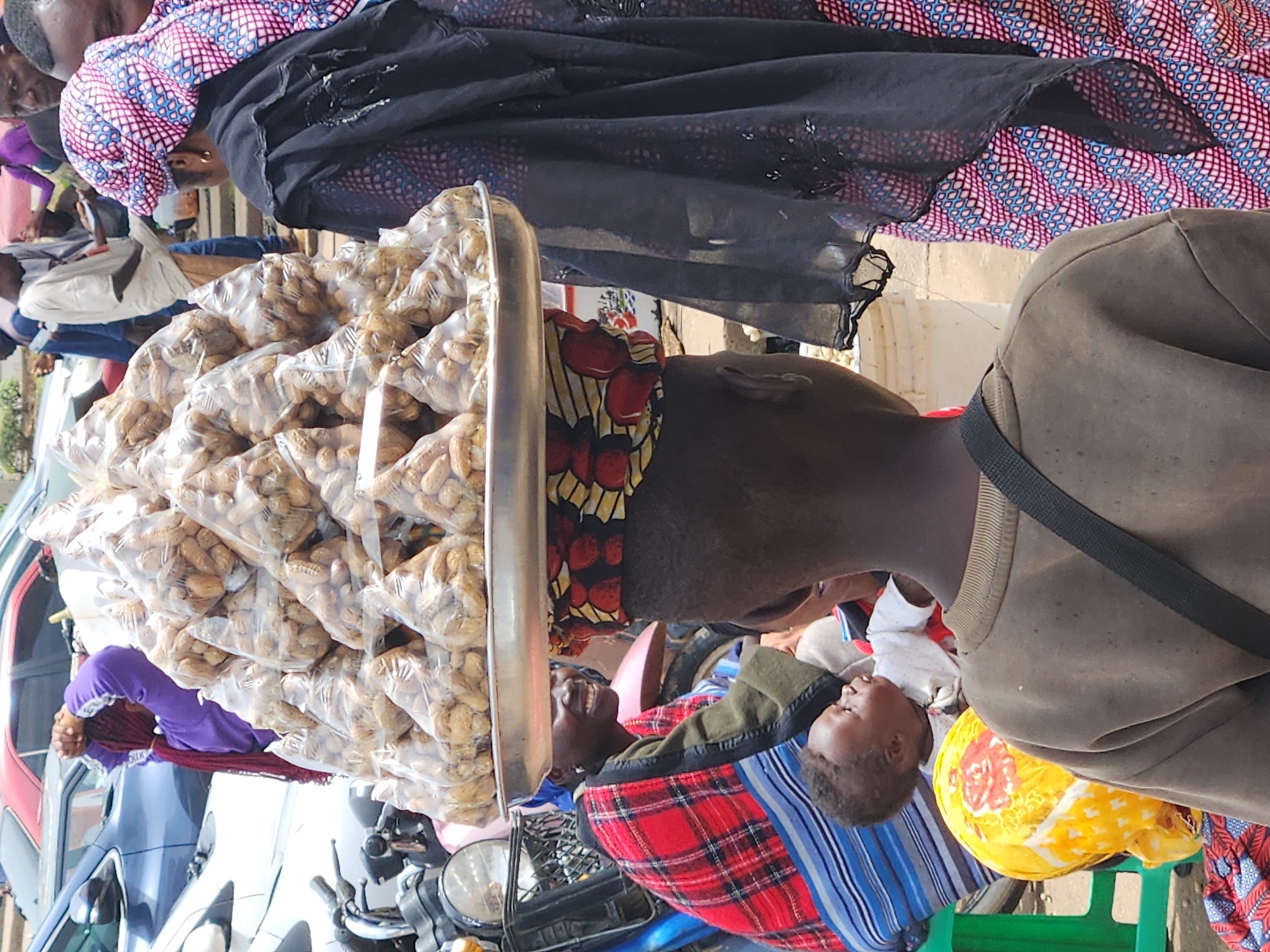 This is a close-up, eye-level, medium shot of a busy street scene in Jos, Nigeria. In the foreground, a young person wearing a brown sweatshirt with a black strap across their chest faces away from the camera. To their right, a woman wearing a black head covering and a patterned blue and pink dress is partially visible. To the left and slightly behind the young person, another woman wearing a red and black checkered blanket-like garment smiles at the camera. She is holding a baby, who is looking upwards. Perched on the woman's head is a large metal tray piled high with many small plastic bags of peanuts. Behind the main subjects, several other people and vehicles, including motorcycles and cars, are visible, suggesting a bustling market or street. The environment appears to be outdoors in a city setting.