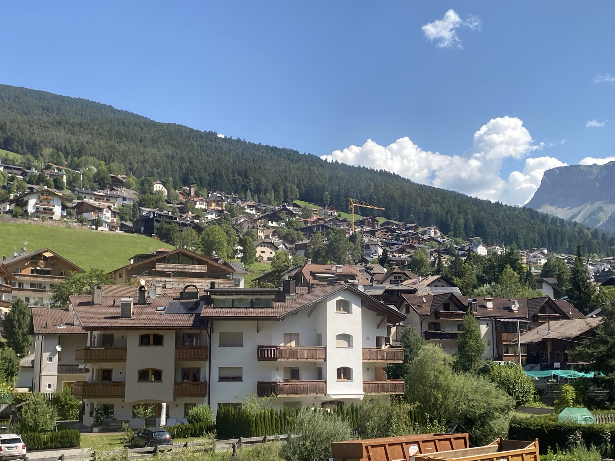 The image depicts a village nestled on a steep, tree-covered mountainside in Kastelruth - Castelrotto, Italy. The architecture is characterized by traditional alpine houses with brown tiled roofs and wooden balconies. Many of the buildings are clustered together, ascending the slope. A bright yellow construction crane is visible amidst the buildings on the upper slope, suggesting ongoing development or renovation. The foreground shows a residential building with multiple stories, balconies, and a well-maintained lawn. Two cars are parked on the left side of the image, one white and one dark grey. The sky is a clear blue with a few scattered white clouds. The overall impression is of a picturesque mountain village bathed in sunlight.