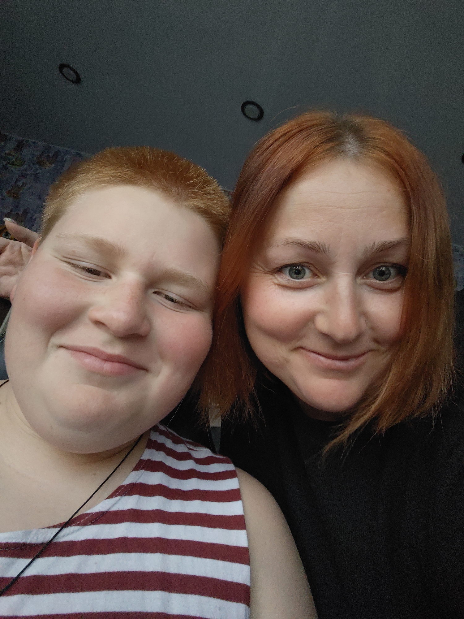 Two individuals, an adult female and a younger person, are posing for a selfie. Both have red hair. The younger person is on the left, with short, spiky hair, and is wearing a red and white striped shirt. The adult female is on the right, with shoulder-length hair, and is wearing a black top. They are both smiling and appear to be indoors, possibly in a home. The background is blurred, but suggests a domestic setting with some wallpaper visible on the left and a ceiling with light fixtures. There is no indication of the location being in Марковский, Russia.