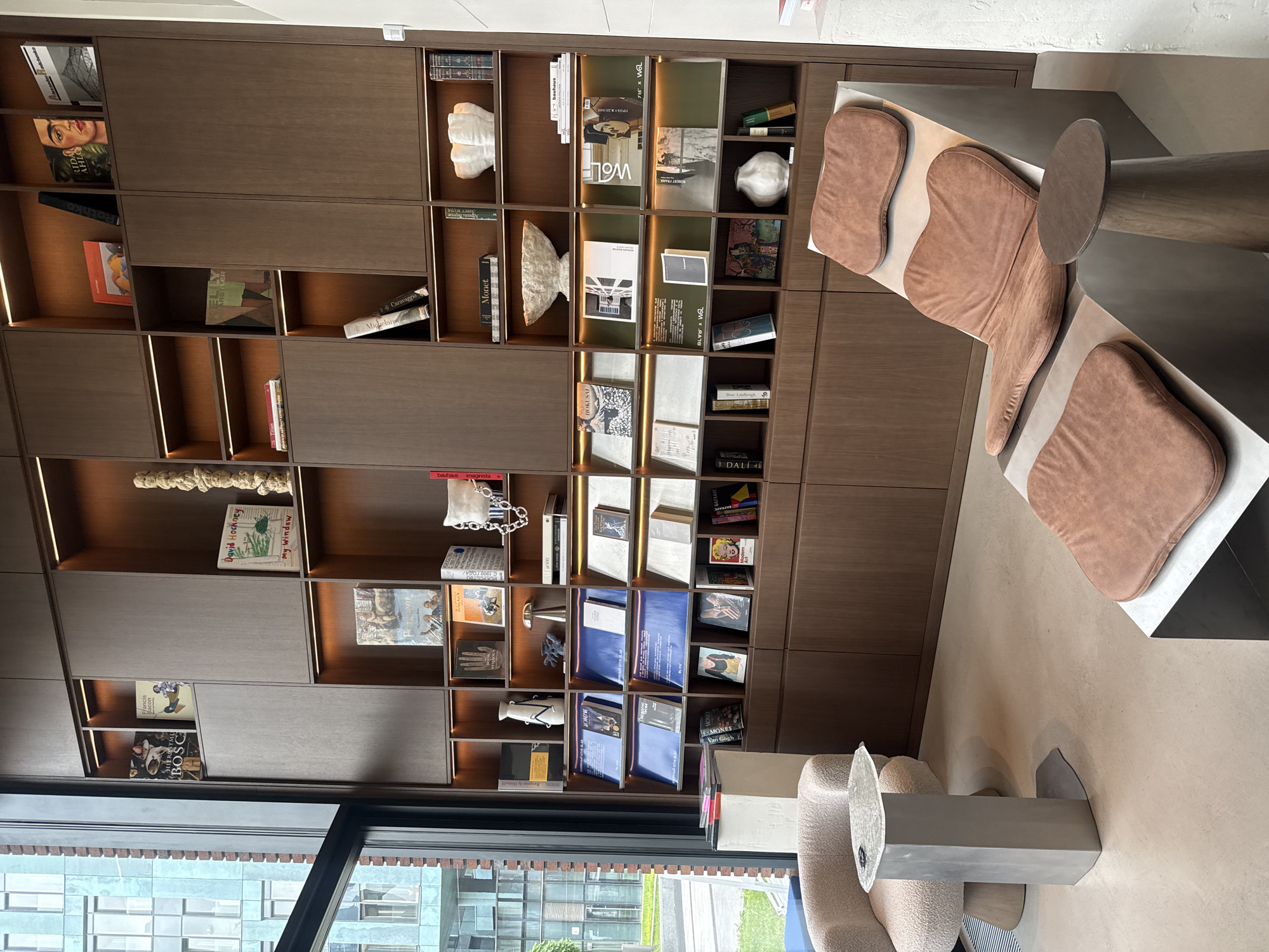 This media file depicts a modern and sophisticated interior space, likely a lounge or common area in an urban setting like Moscow, Russia. The dominant feature is a large, dark wooden built-in shelving unit, combining open shelves with closed panels.
The shelves are meticulously arranged with a variety of books, predominantly art books and monographs, alongside decorative objects. Visible book titles and authors include "HIERONYMUS BOSCH", "Francis Bacon", "David Hockney - My Window", "Monet", "Van Gogh", "Caravaggio", "Rothko", "HOKUSAI", "DALI", and "Dur Lielbergh". Other textual elements observed are "bauhaus imaginista", "Miguelary", "ELLY", "ARSTLER", "WOLF", "BLVB X WO", and the Cyrillic word "ДОВЕРИЕ" (Trust). Interspersed among the books are various sculptures and vases, such as white ceramic pieces resembling abstract forms or corals. The shelves feature integrated LED strip lighting, highlighting the displayed items.
In the right and foreground of the image, there is a contemporary seating area. It consists of a long, low table or platform with an irregular, organic shape, topped with several brown upholstered cushions that serve as individual seats. A small, round side table with a thick, cylindrical base is positioned next to this seating unit. Further to the bottom right, another light-colored, textured upholstered armchair and a geometric side table holding a clear, sculptural object are partially visible.
A large window on the left side of the room offers a view of modern architecture, featuring glass-fronted buildings and some greenery, indicating a daytime setting. The overall atmosphere is calm and aesthetically curated, emphasizing contemporary design and cultural interests.