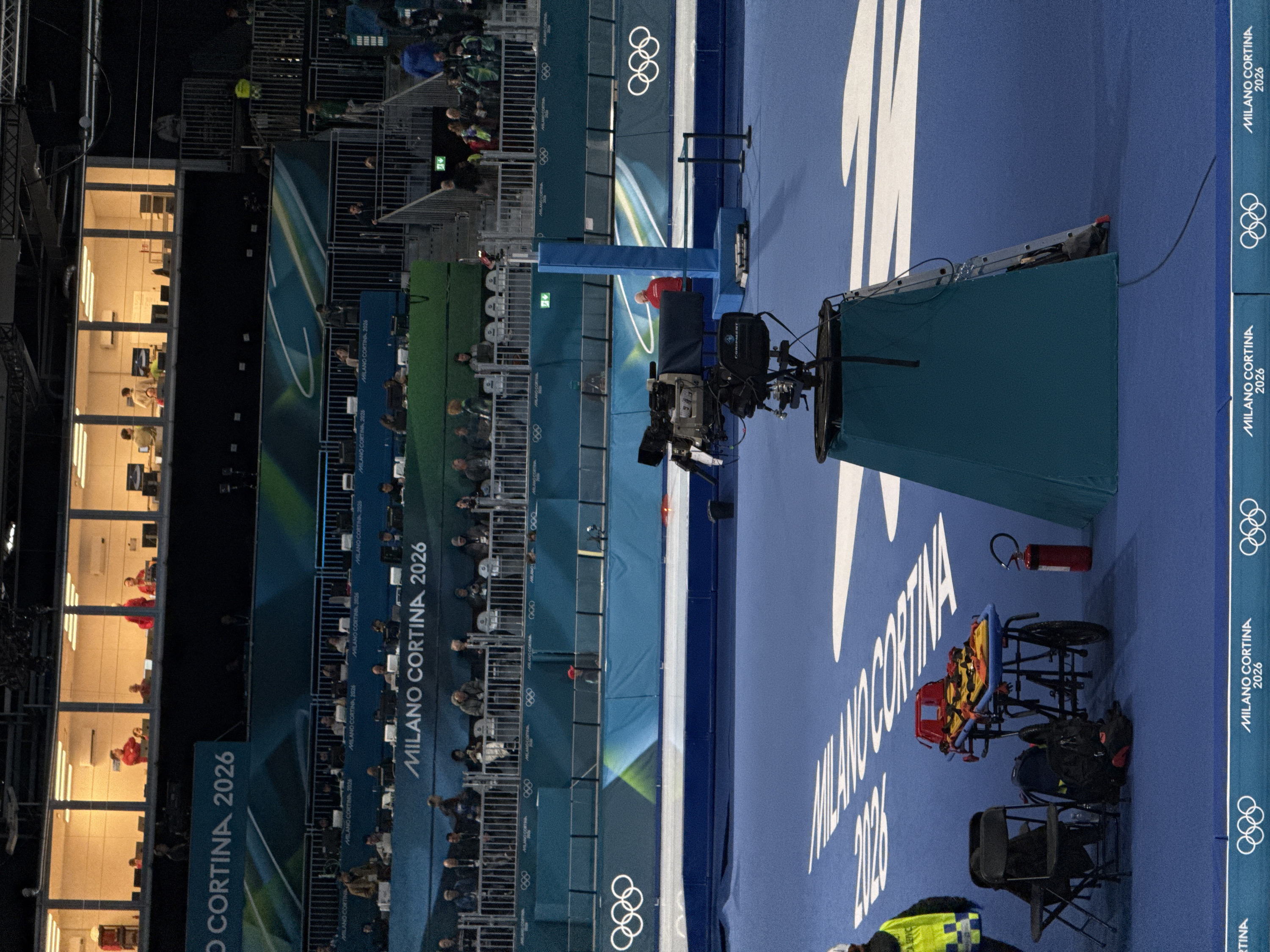 This image captures an indoor sports arena or event venue, primarily focusing on broadcast equipment and a section of spectator seating, with strong branding for "MILANO CORTINA 2026." The image is oriented vertically, showing the scene from a slightly elevated perspective looking down and across the venue.
In the mid-ground, a prominent professional broadcast camera is positioned on a tripod, partially covered by a dark green fabric. To the right of the camera, a blue floor or wall displays large white graphics and text, including "MILANO CORTINA 2026." Further to the right, a red fire extinguisher and a yellow and red medical stretcher or cart are visible on the blue surface. A ladder is leaning against a raised platform near the camera.
The left side of the image features multiple tiers of stadium-style seating, largely occupied by spectators or event personnel. These seating areas are structured with metal railings. Below these stands, a long, rectangular room with large windows is illuminated from within. Inside this room, several individuals are seated at desks, each with multiple computer monitors, suggesting a control room, media center, or broadcast operations hub. Some individuals in this room are wearing red or light-colored shirts.
Throughout the venue, various blue and green banners and hoardings display the text "MILANO CORTINA 2026" and the iconic Olympic rings logo, indicating the event is related to the Milano Cortina 2026 Winter Olympics and Paralympics. The overall lighting is artificial, suggesting an indoor setting. The scene appears to be either during an ongoing event, or during setup/rehearsal, given the presence of people, equipment, and fully branded environment.