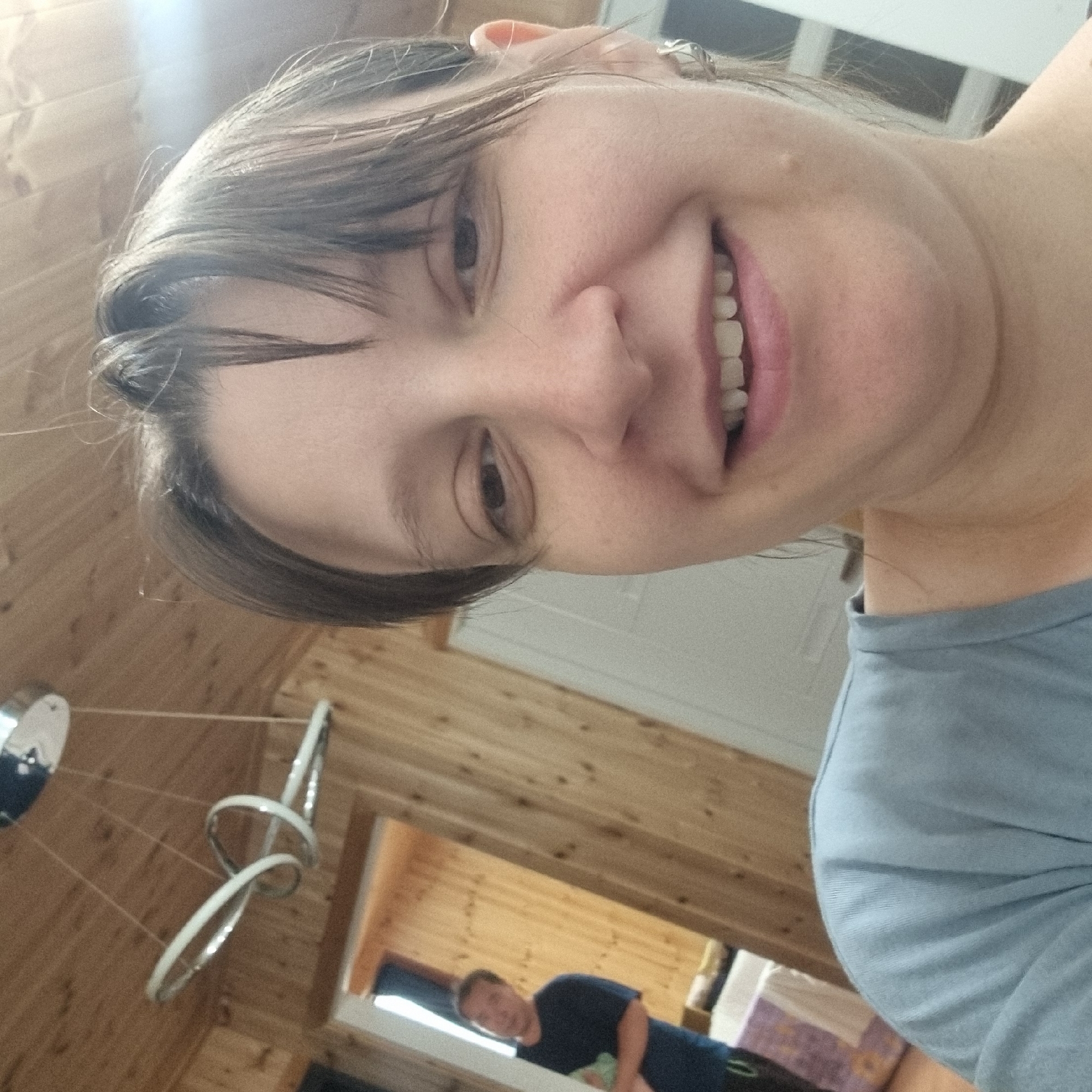 The image captures a selfie taken indoors, likely in a residential setting with wood-paneled walls and ceiling. In the foreground, a woman's face is prominently featured, with her right eye and mouth visible. She has dark hair and is wearing a light blue shirt. Her expression appears to be a smile. In the background, through an open doorway, a man wearing a dark blue t-shirt is visible, standing and looking towards the camera. He is in a room with wooden walls and what appears to be a bed or seating area in the lower left corner. A modern-looking light fixture hangs from the ceiling. The overall impression is a casual, indoor snapshot. The location is specified as Dunnae-myeon, South Korea.