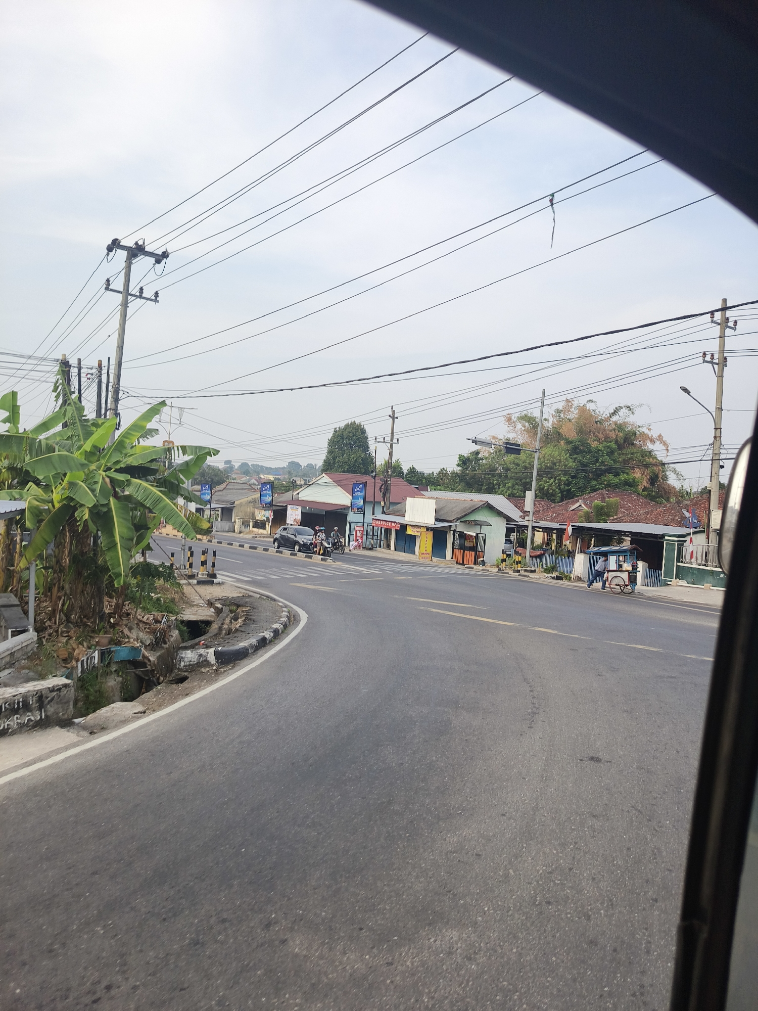 This is a street-level, outdoor, daytime, medium shot captured from a vehicle looking out towards a two-lane asphalt road. The road curves to the right and is bordered by a curb and a drainage ditch on the left, and various small businesses and residential buildings on the right. Power lines are strung across the sky, supported by utility poles.
On the left side of the road, there are lush green banana trees. Further down the road, several cars and motorcycles are visible, suggesting moderate traffic. Some of these vehicles appear to be parked or in motion near the storefronts.
The buildings on the right side of the road are single-story structures with tiled roofs, painted in various colors, including white, blue, and orange. Some of these buildings have signage, indicating they are shops or businesses. A person is seen pushing a cart along the sidewalk. The sky is overcast with a pale, hazy quality.
The image is believed to be from the city of Pangkalpinang, Indonesia, as evidenced by the presence of common Indonesian street scenes, including the architecture, vegetation, and road infrastructure. The scene depicts a typical commercial and residential area in the city.