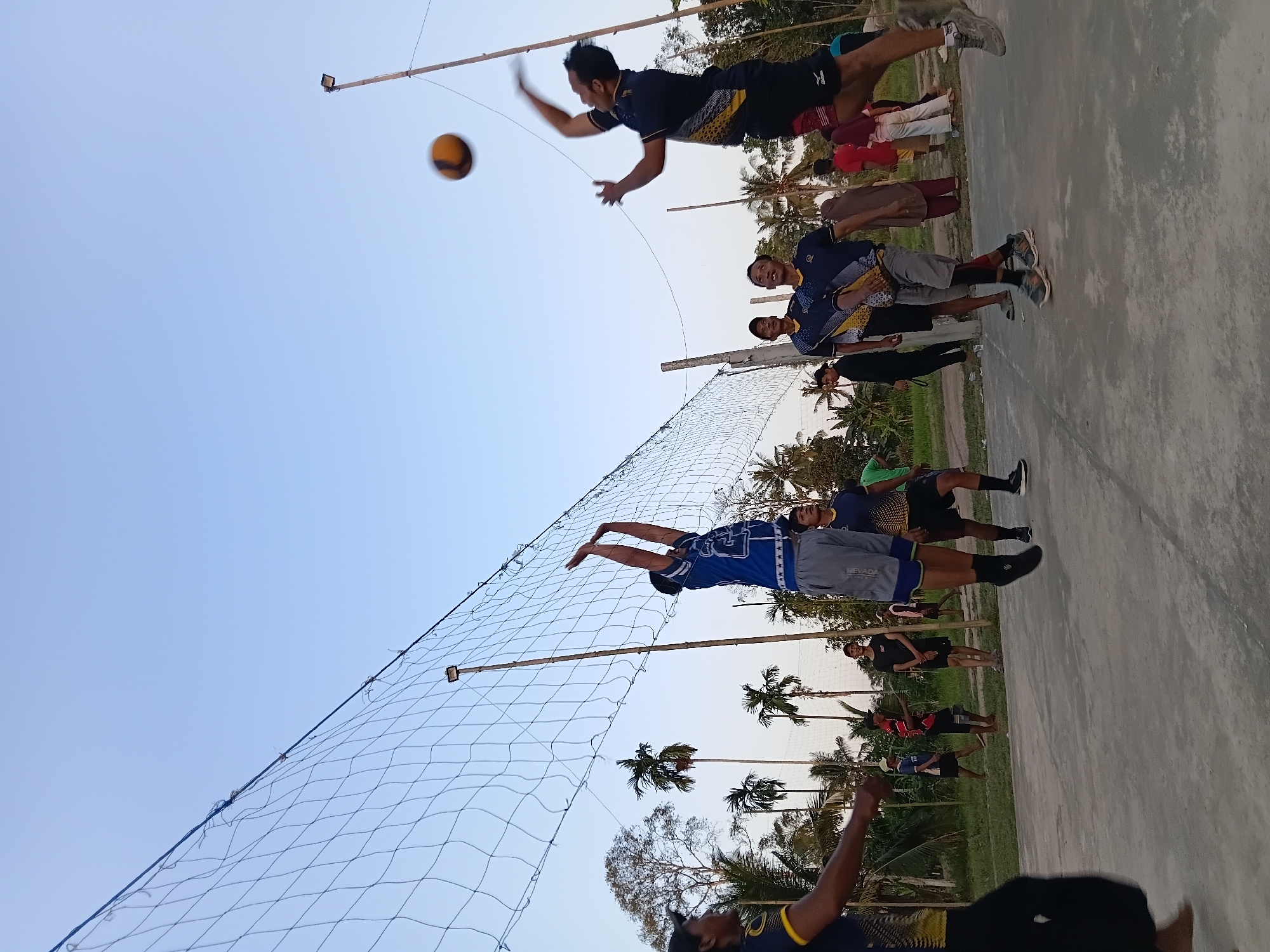 In the city of Margo Rukun, Indonesia, a group of men are playing volleyball on an outdoor court. The court appears to be made of concrete. The sky is clear and the time of day is likely late afternoon or early evening. A volleyball net, made of blue netting and supported by wooden poles, divides the court.

Several men are actively engaged in the game. Two players are in mid-air, one jumping to spike the ball and another blocking. The player spiking the ball wears a dark blue t-shirt with yellow accents and black shorts. The player attempting to block wears a blue and white t-shirt with gray shorts. Other players are positioned on the court, some watching the action and others preparing to receive the ball. One player on the left side of the image is partially visible, wearing a dark shirt with yellow accents and black pants.

In the background, there is lush vegetation, including palm trees, suggesting a tropical or subtropical environment. A few spectators are present on the sidelines, some standing and others appearing to be seated. A light pole with a lamp is visible near the net on the right side of the court. The overall scene depicts a casual game of volleyball in an outdoor setting.