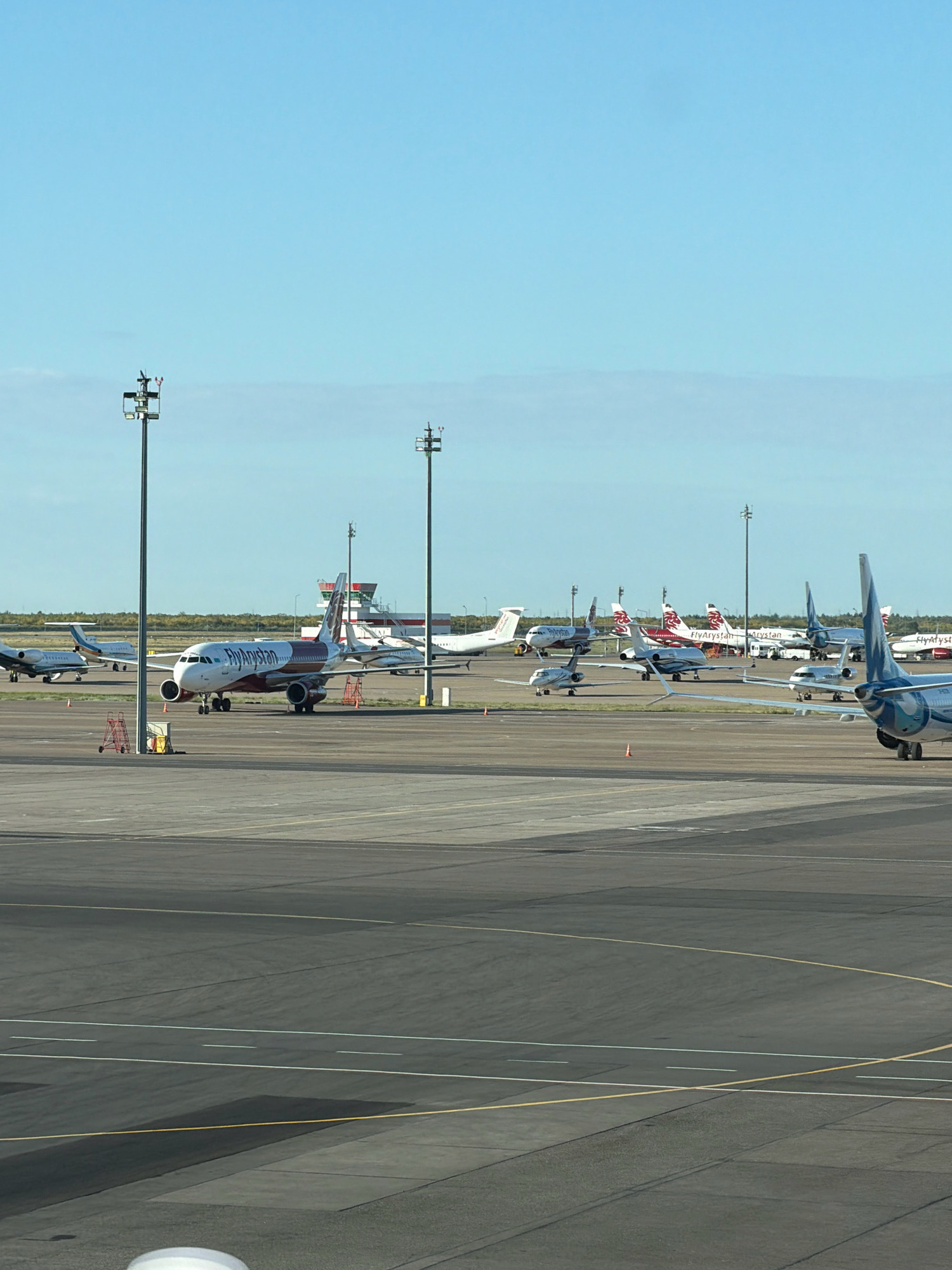 An aerial view of Astana International Airport in Astana, Kazakhstan, shows an tarmac filled with airplanes. The airport features a mix of commercial and private jets parked on the runway and taxiway. A central terminal building with a distinctive control tower is visible in the background. The sky is clear blue with a few wispy clouds.