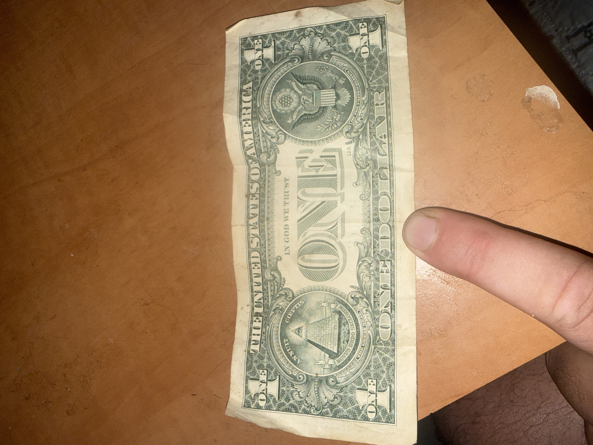 A person's thumb is visible in the foreground, obscuring the bottom right corner of a crumpled one-dollar bill. The bill lies on a textured, reddish-brown surface that appears to be wood. The bill's obverse side is predominantly visible, showing the Great Seal of the United States on the right and the Eye of Providence pyramid on the left. The words "THE UNITED STATES OF AMERICA" and "IN GOD WE TRUST" are clearly legible. The bill is creased and slightly worn. The background is out of focus and consists of the same reddish-brown surface. There is no information to suggest this image is located in Herzliya, Israel.