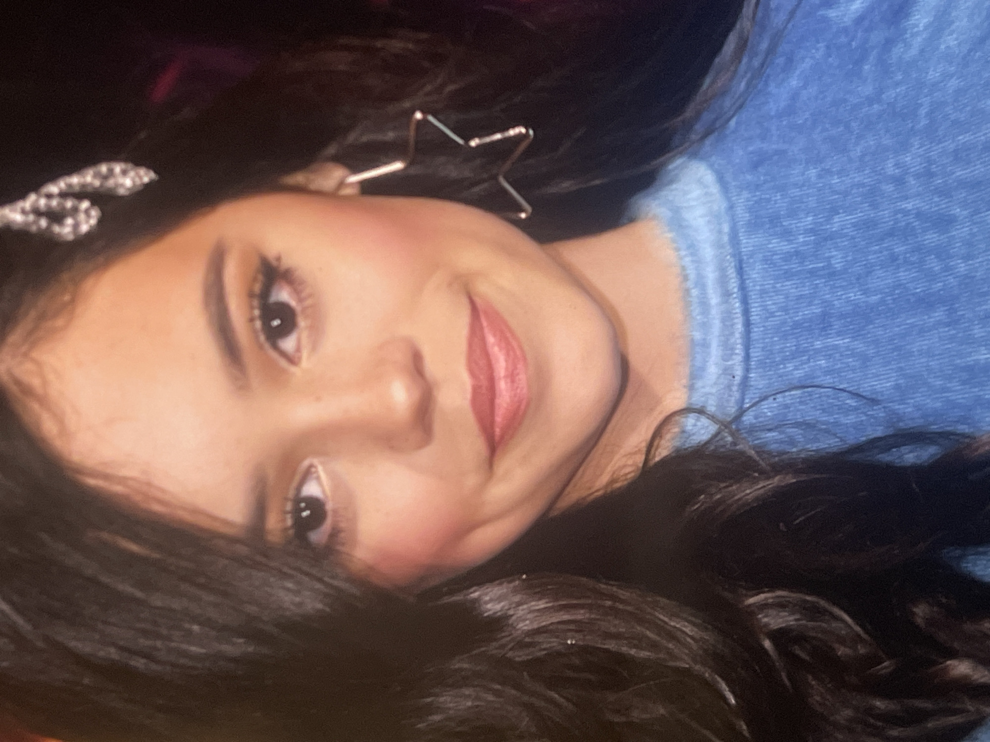 A young girl with long, dark, wavy hair is captured in a close-up, rotated portrait, appearing to be lying down. She has light skin, dark eyes, and a soft, closed-mouth smile. Her cheeks show a rosy blush, and her lips are adorned with pink lipstick.
She wears two distinct accessories: a silver, shimmering hair clip with an intertwined, possibly jeweled, design is visible in her hair on the left side of the frame. On her right ear, she has a prominent gold-toned earring shaped like an open five-pointed star. Her upper body is covered by a blue, textured garment, resembling a sweatshirt or sweater, with visible stitching details.
The background is dark and out of focus, suggesting an indoor setting with attention drawn to the subject. The lighting highlights her face, indicating an intentional photograph. The girl's expression conveys a sense of pleasantness and contentment. There are no specific location cues within the image itself that denote Poltava, Ukraine, and no visible text.