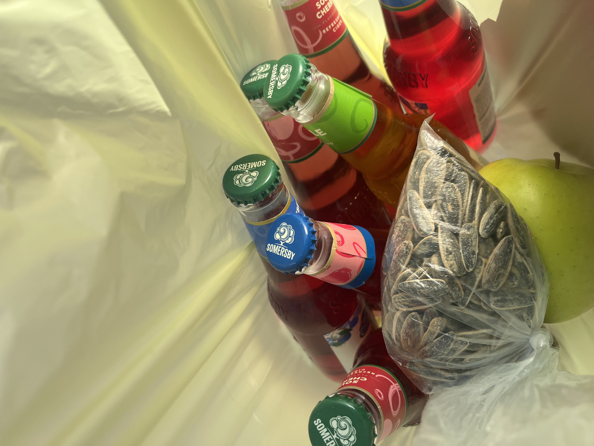 The image shows a plastic bag containing several items. There are multiple bottles of Somersby cider with different flavors: cherry, apple, and potentially others based on the cap colors. A bag of sunflower seeds and a green apple are also present. The bag is light yellow, suggesting it is a plastic shopping bag. The background and lighting suggest an indoor setting. The city of Lozarevo, Bulgaria is not identifiable in the image, so I am unable to verify or make any inferences about this being in Lozarevo.