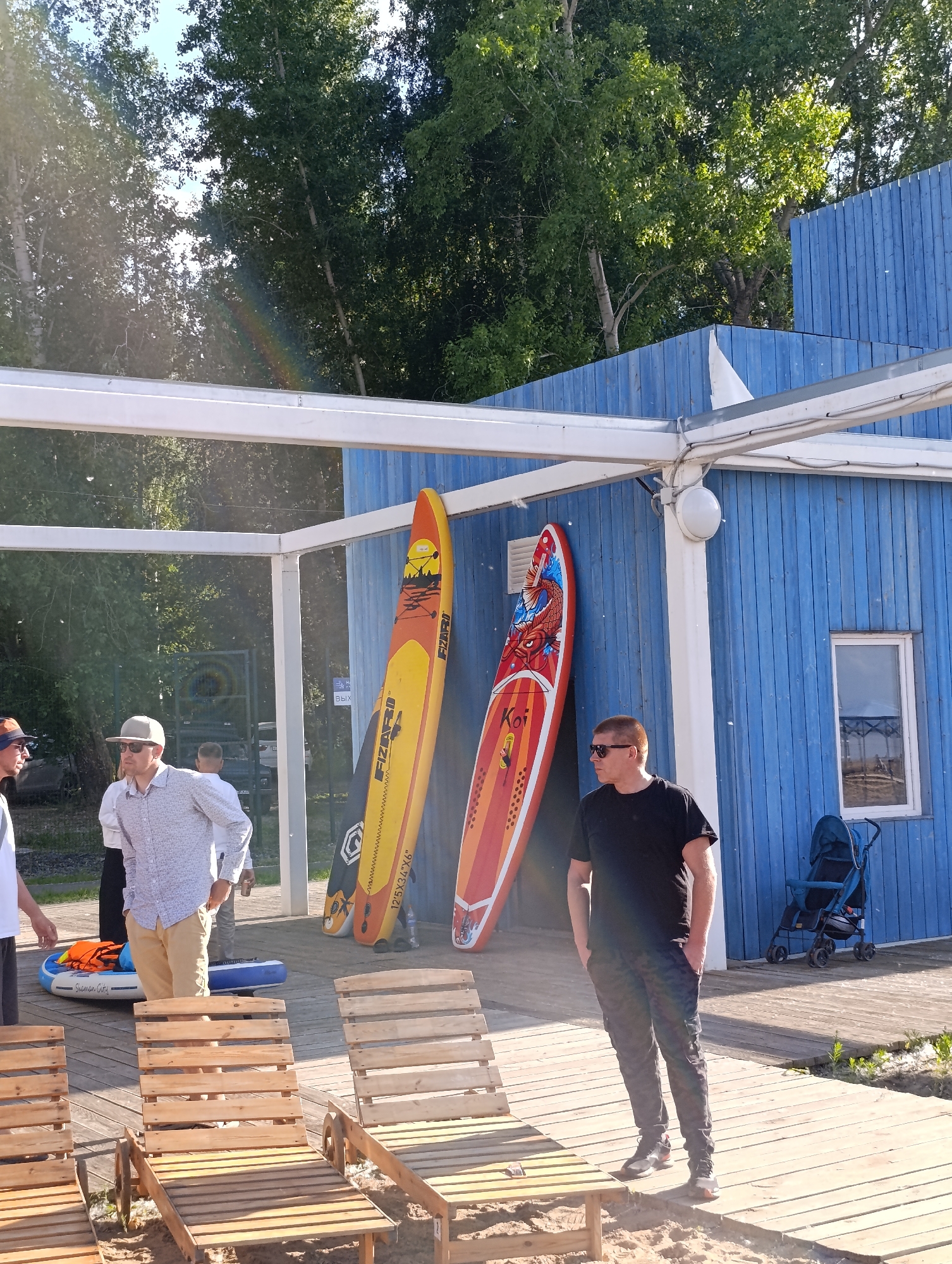 A group of people are gathered outdoors near a blue wooden building with a white awning. Two colorful paddleboards are propped against the building. In the foreground, several wooden lounge chairs are set up on a boardwalk. A man in a black t-shirt and patterned pants stands with his hands in his pockets, looking to the side. Another man in a patterned shirt and cap is looking down. In the background, trees are visible, and a stroller is parked near the building. The scene appears to be a recreational area, possibly a beach or lakeside resort.
