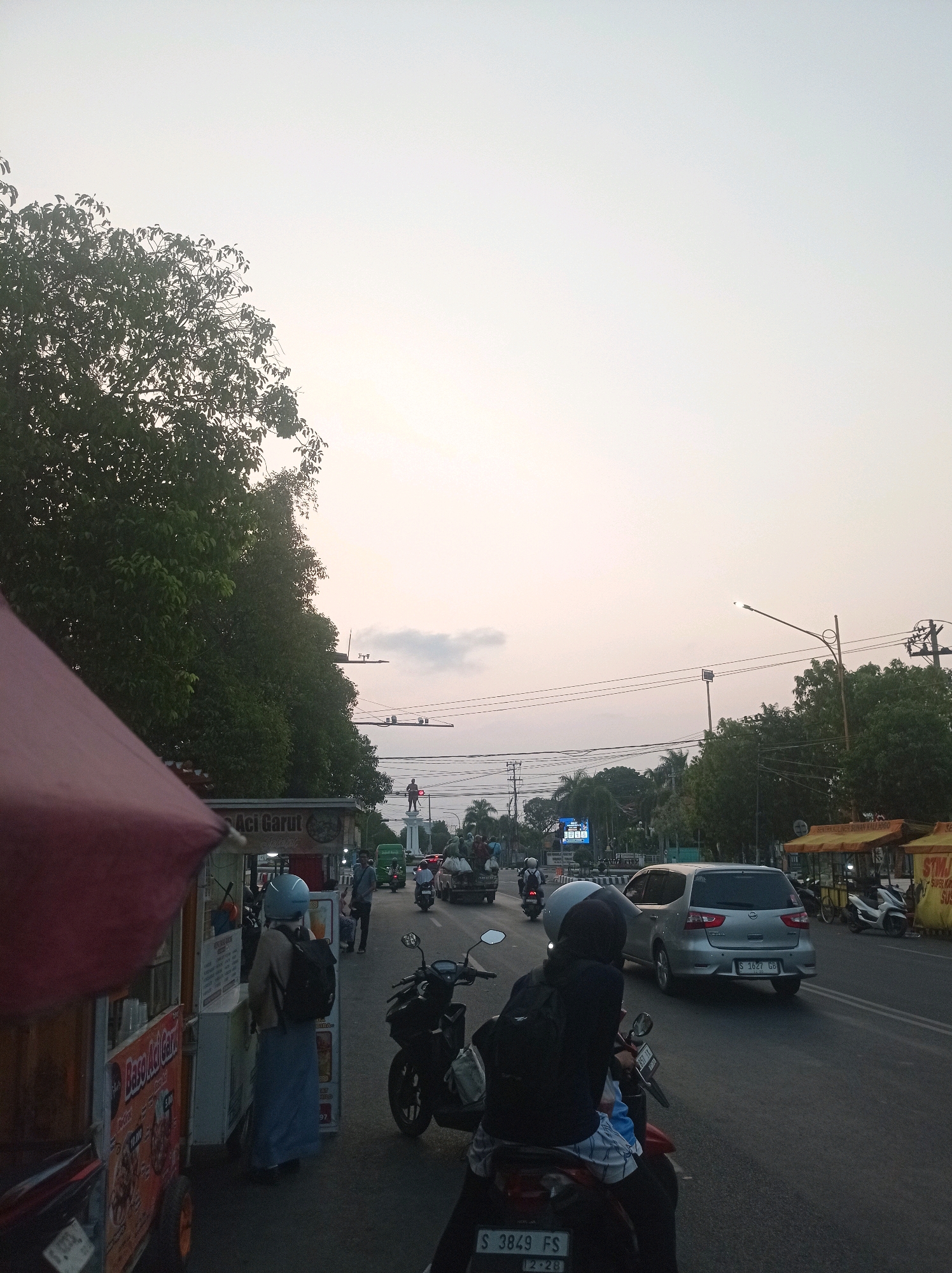 The image depicts a street scene in Tuban, Indonesia, likely during dusk or early evening, as indicated by the soft, diffused light and the sky's muted tones. The vantage point is from the side of a street where several food carts are set up. In the foreground, a food cart with a red awning is partially visible on the left. Next to it, another food cart displays the text "Baso Aci Garut" and menu prices. A person wearing a light blue hijab and a dark backpack stands facing away from the camera, looking at the food cart.
Further down the street, there is a moderate amount of traffic, including motorcycles and cars. A silver minivan with license plate S 1627 GB is moving away from the camera. In the distance, a statue, possibly a monument, stands prominently on a white pedestal, with traffic lights and power lines visible in the background. Trees line the sides of the street, and a few more food stalls are set up on the right side, one with yellow signage that reads "STIM..."
Several individuals are present, some on motorcycles, others walking. The overall atmosphere suggests a typical evening in a busy urban area. The sky is a pale, hazy expanse, with subtle gradients of light blue and white, hinting at the fading daylight.