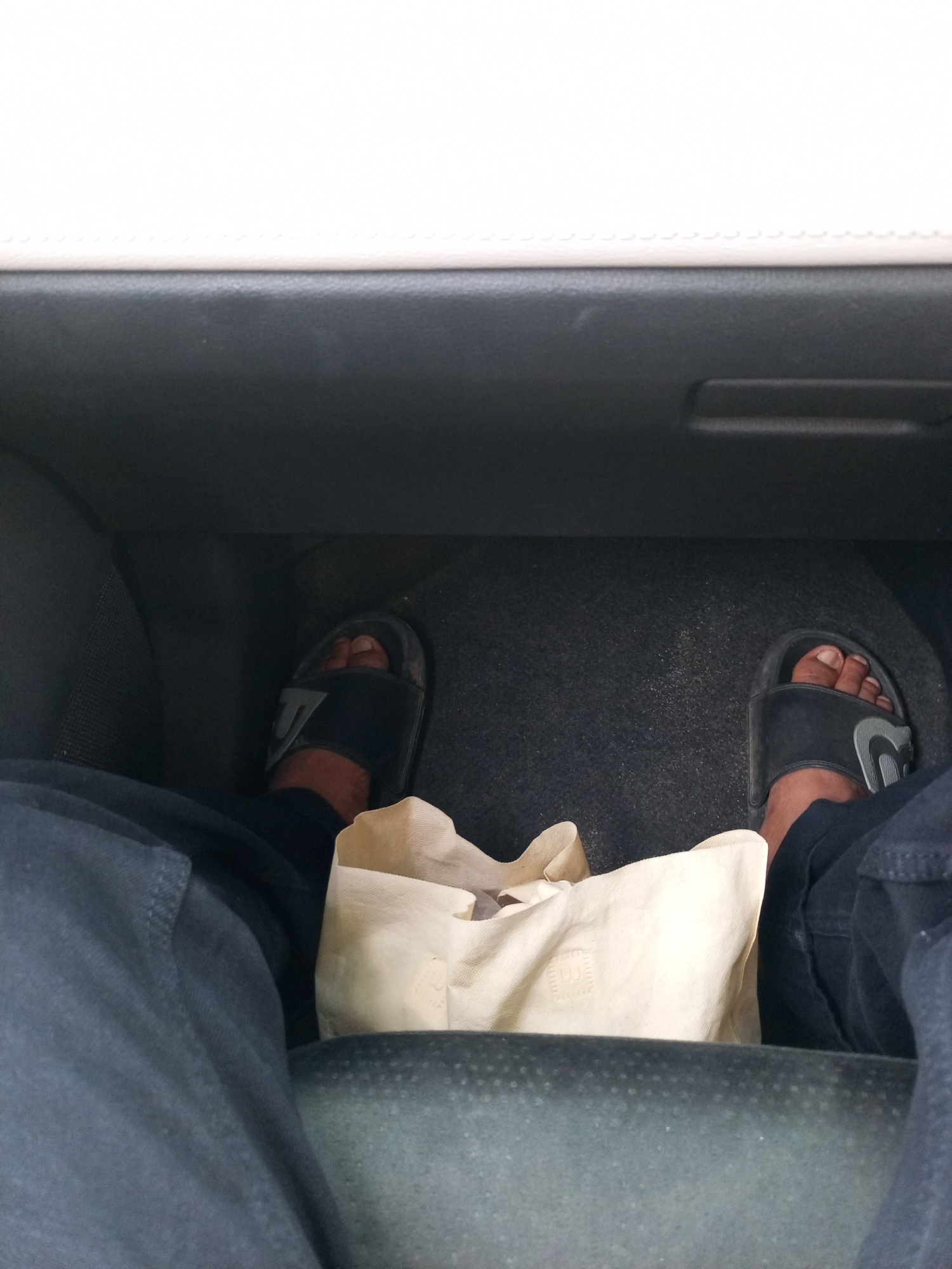 The image captures a low-angle view of a person's feet and legs inside a vehicle, likely a car, as indicated by the dashboard and seating material. The individual is wearing black slides with a grey Nike logo on the left foot, and a similar black slide on the right foot. Dark blue pants are visible, covering the legs down to the ankles. Between the feet, a beige fabric bag is placed on the floor. The floor of the vehicle is dark grey and appears to have some dust or debris. The background shows the interior of a car, with a black dashboard and a cream-colored upper trim. The specific location in Lhokseumawe, Indonesia, is not discernible from the image.