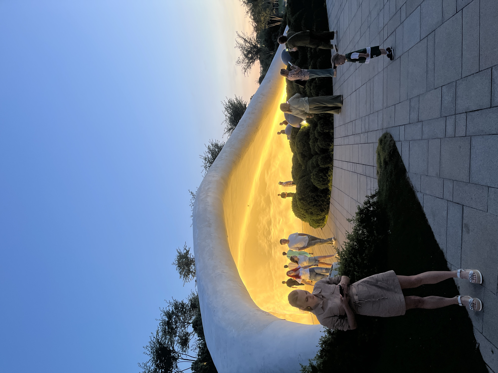 The image depicts a contemporary outdoor art installation or architectural feature in Krasnodar, Russia, likely at a park or public space. The structure is a large, white, curved form with an interior illuminated by a warm, golden light. The time of day appears to be dusk, with a clear blue sky transitioning to darker tones.
Several people are present, interacting with the space in various ways. In the foreground on the left, a woman wearing a light-colored dress and platform sandals is looking at her phone. Behind her, a group of people are walking and observing the installation. Further back, a child in a green and white outfit is standing on the paved walkway.
The ground is paved with large, light-colored square tiles, with patches of green grass and dark, manicured bushes surrounding the illuminated structure. Trees with dark foliage are visible in the background against the twilight sky. The overall atmosphere suggests a public gathering place designed for leisure and appreciation of art or architecture.