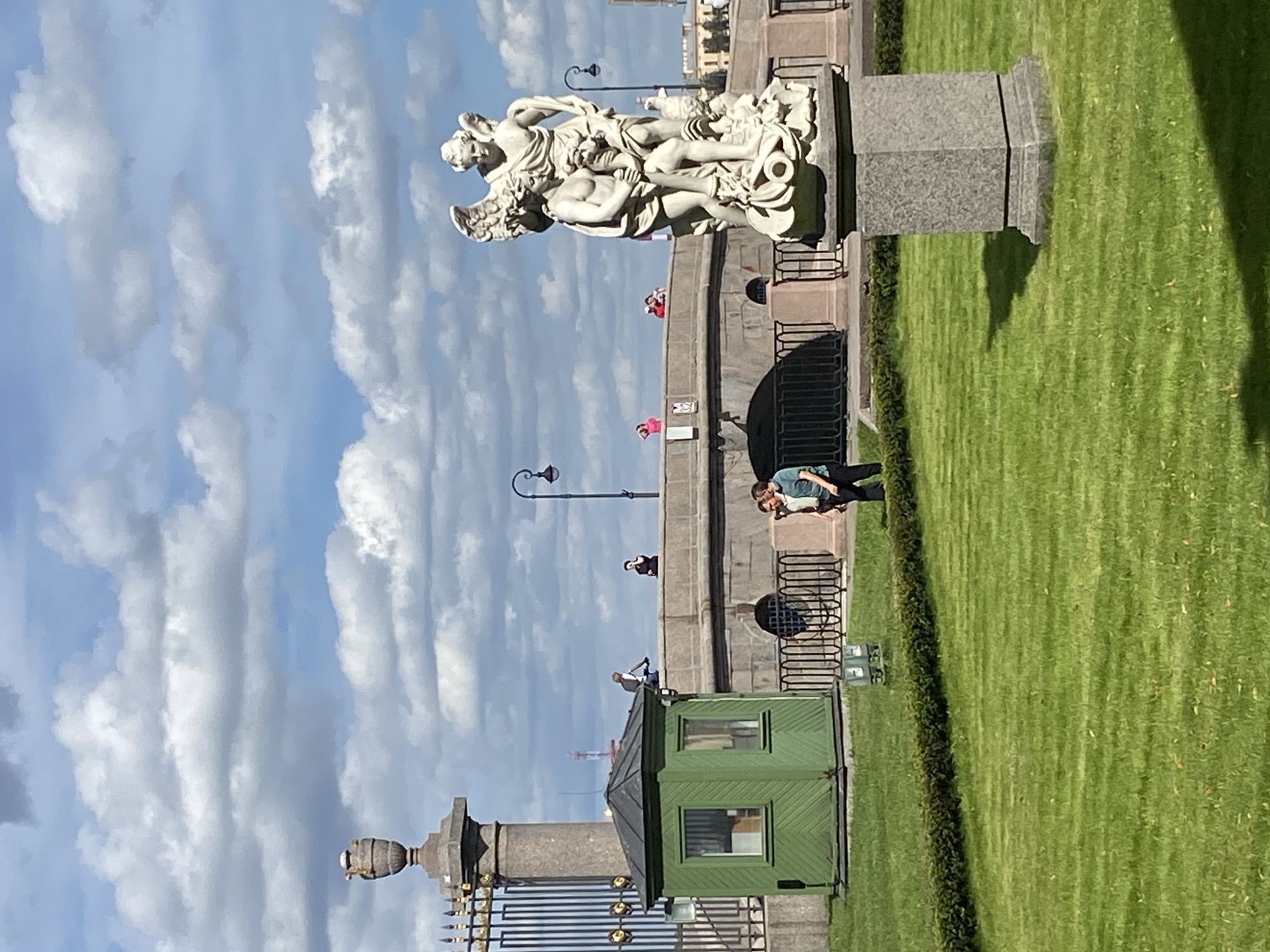The image depicts a scene in Saint Petersburg, Russia, likely near the Neva River. In the foreground, a meticulously manicured green lawn is cut by a low, dense hedge. To the right, a stone pedestal supports a white marble statue of two figures, appearing to be cherubs or putti, intertwined. Behind the lawn, a low, stone bridge with arched openings and ornate black railings spans a waterway.
On the bridge, several individuals are visible. Two men are standing in conversation on the far side, near a lamp post. Further along the bridge, a person is seated on the railing, seemingly riding a bicycle, while another individual stands nearby. A few more people are scattered along the bridge's length.
To the left of the image, a small, hexagonal green wooden structure with two windows stands on the grass. Behind this structure, a tall, granite pillar topped with an urn is part of a decorative metal fence. The background features a bright blue sky with scattered white clouds. The overall scene suggests a pleasant day in a park or public space within the city.