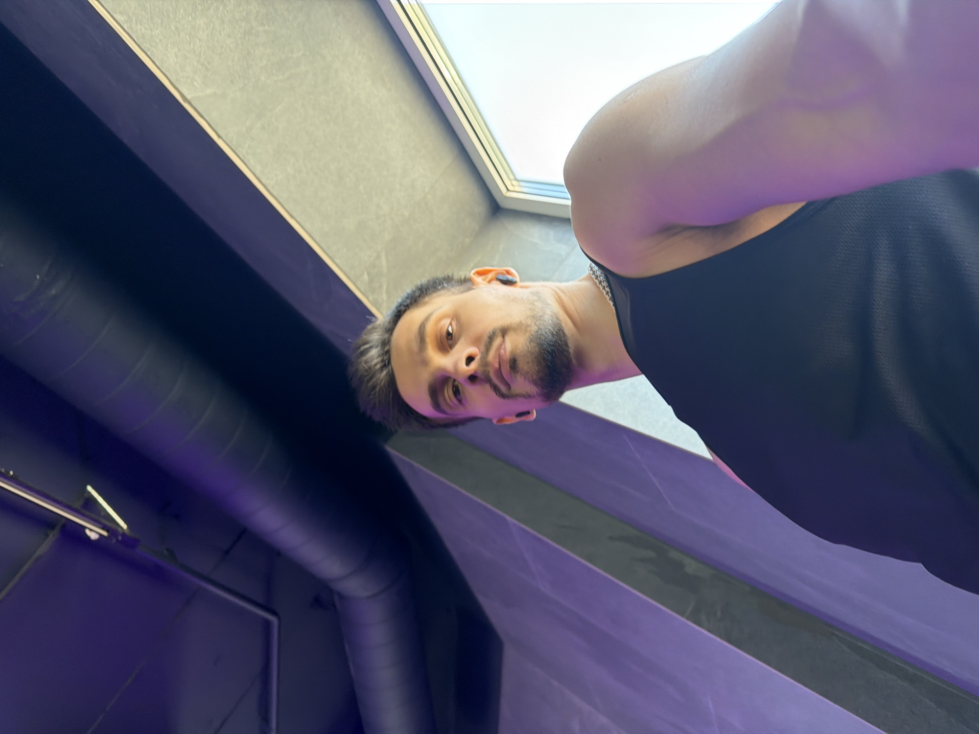 A low-angle, selfie-style shot shows a person with dark hair and stubble looking down at the camera. They are wearing a black tank top and a chain necklace. Earbuds are visible in their ears. The background features a window with a bright sky, a textured wall with purple lighting, and industrial-looking pipes and fixtures on the ceiling. The image appears to be taken indoors, possibly in a gym or studio. The exact location within Krasnoyarsk, Russia, cannot be determined from the image alone.