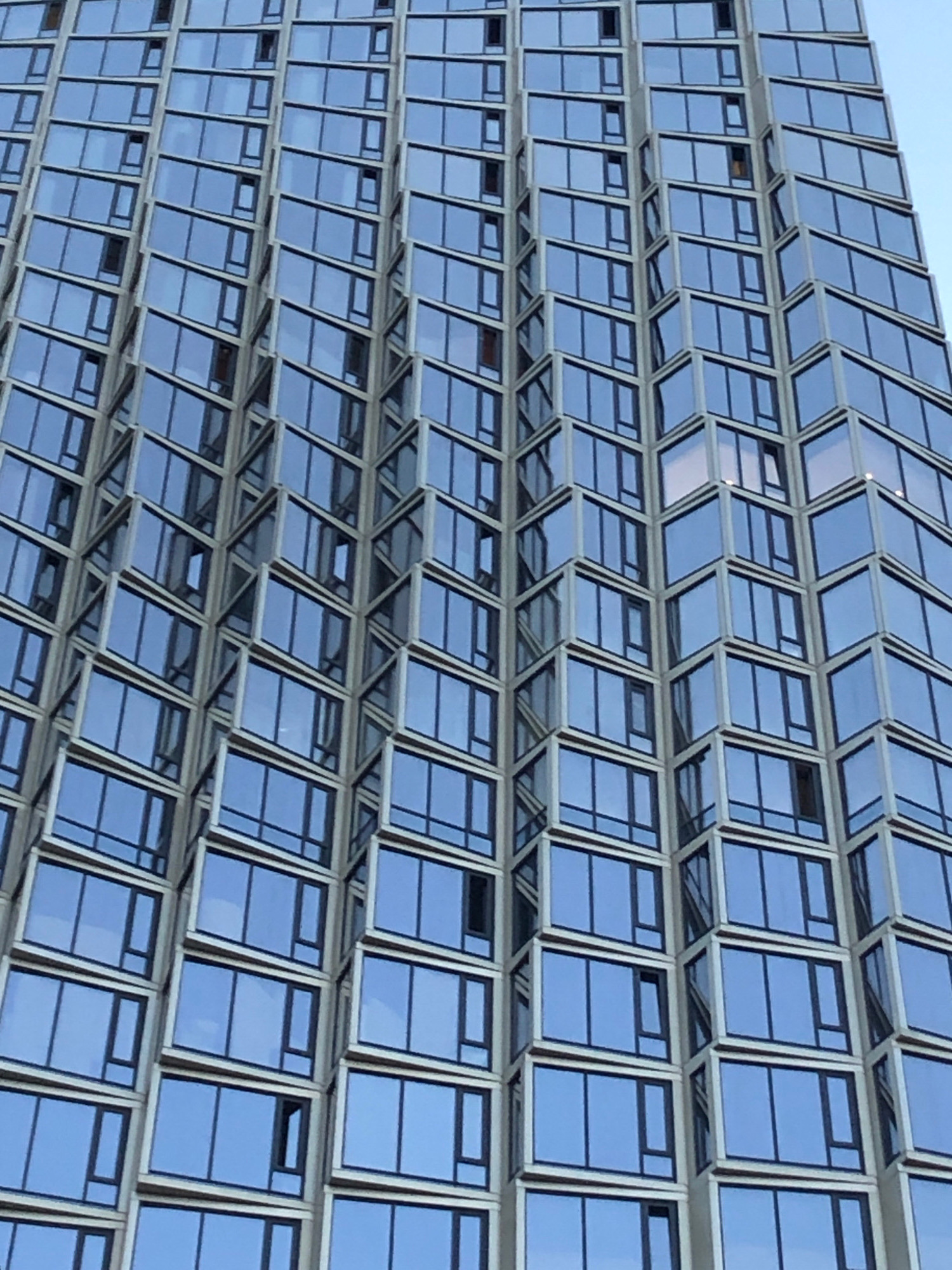 The image shows a close-up, low-angle view of a modern skyscraper with a facade of blue glass windows and light-colored framing. The windows are arranged in a staggered pattern, with some protruding outward, creating a textured and dynamic appearance. The building appears to be tall and is reflected in the glass panes, along with a clear blue sky. There are no people or any other discernible objects or activities visible in the image. The architecture suggests an urban environment.