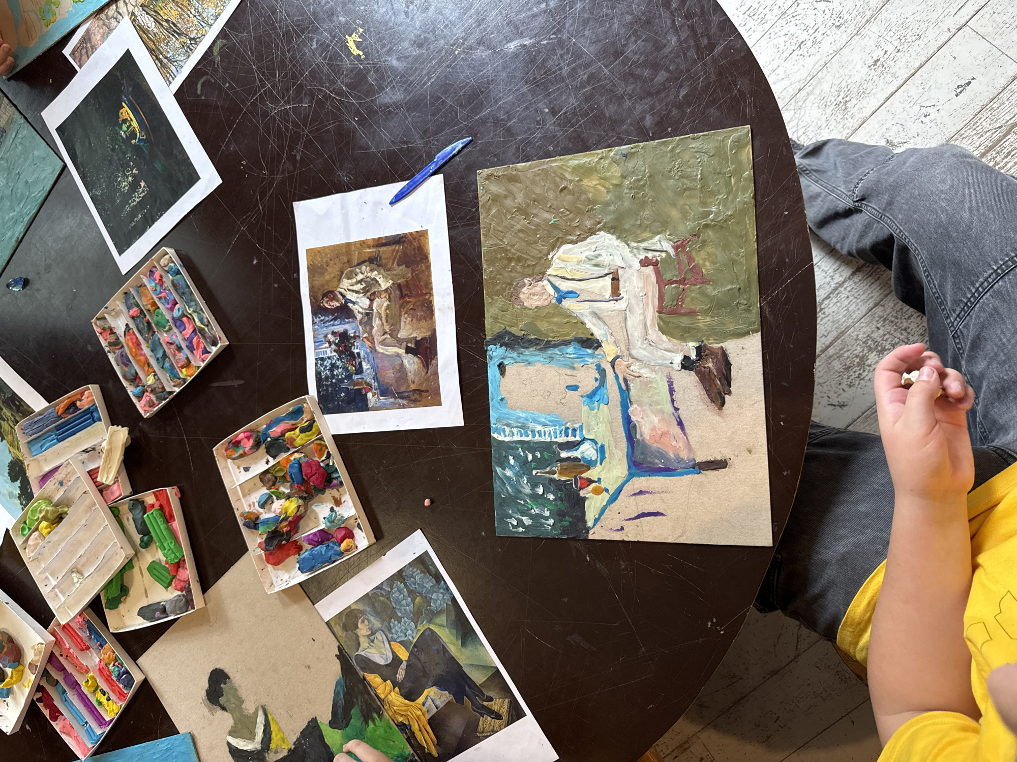 The image shows a person's hands and legs from an overhead perspective, resting on a dark, circular table. The table is covered with various art supplies, including colorful modeling clay in partitioned boxes, small canvases, and printed art reproductions. One painting depicts a figure in a white suit sitting on a chair, while another shows a still life with bottles and fruit on a table. Another reproduction shows a scene with a boat on dark water. The person is wearing a yellow shirt and dark jeans. The scene appears to be a creative workspace, possibly a studio or a classroom, with art in progress and materials readily available. The flooring beneath the table is light-colored wood, possibly painted or distressed. The overall impression is one of artistic activity and a focus on the creation of art. The location is not specified within the image or its context.