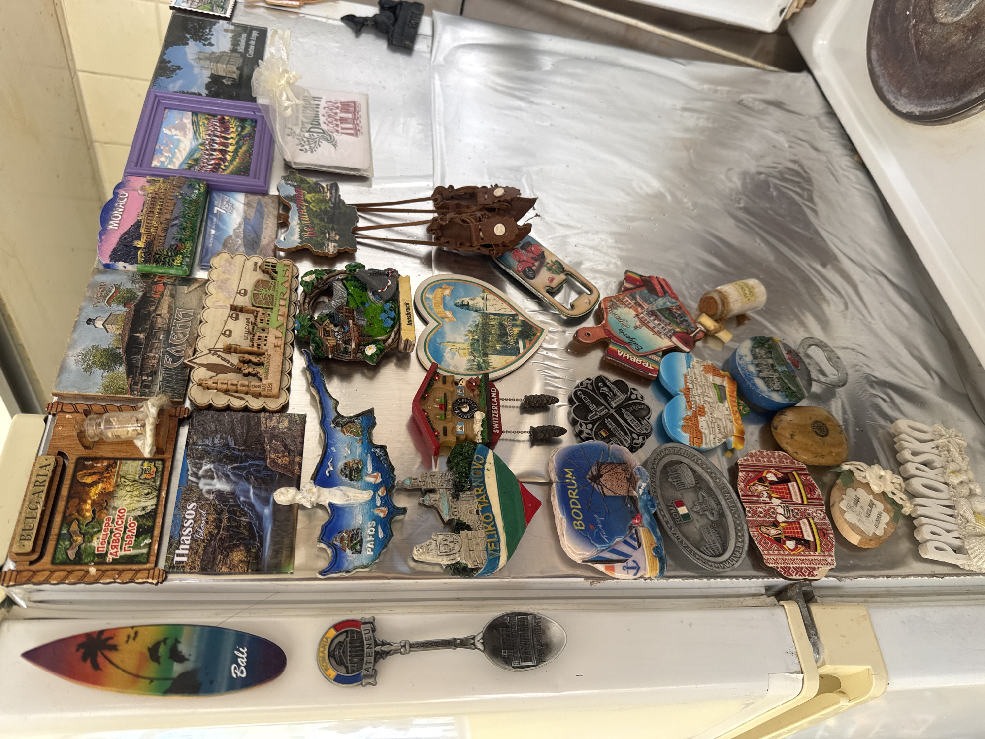 The image shows a collection of souvenir magnets arranged on a metal surface, likely the top of a refrigerator or appliance. The magnets depict various locations, landmarks, and themes from different countries and regions. Some of the magnets include names of places, such as Bali, Romania, Bulgaria, Thassos Island, Pafos, Veliko Tarnovo, Bodrum, Roma, Monaco, and Switzerland. Other magnets feature images of landmarks, landscapes, or cultural symbols. The magnets are of various shapes, sizes, and materials. The surface beneath the magnets appears to be metallic and reflective. There is nothing in the image to connect it to the city of Varna, Bulgaria.