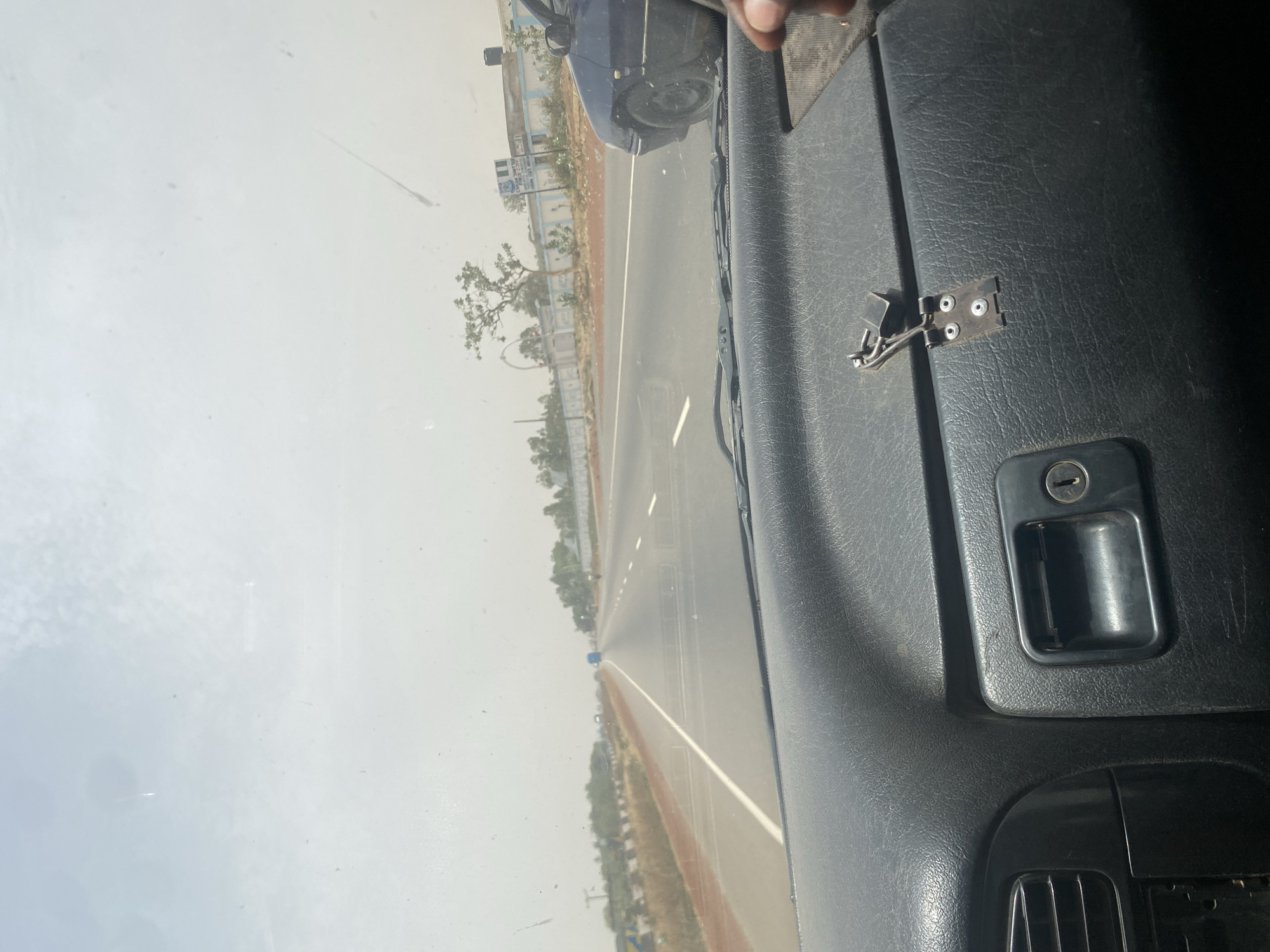 The image depicts a view from inside a vehicle, likely a car or truck, traveling on a highway. The road is paved and has lane markings. The surrounding environment appears dry and dusty, with trees and vegetation visible along the roadside. In the foreground, part of the vehicle's interior is visible, including the dashboard with a latch and a handle, suggesting the presence of a storage compartment. A passing car can be seen to the left. It is challenging to ascertain the precise location as Zangon Aya, Nigeria, with certainty from this image, but the environmental conditions and vegetation are consistent with that region.