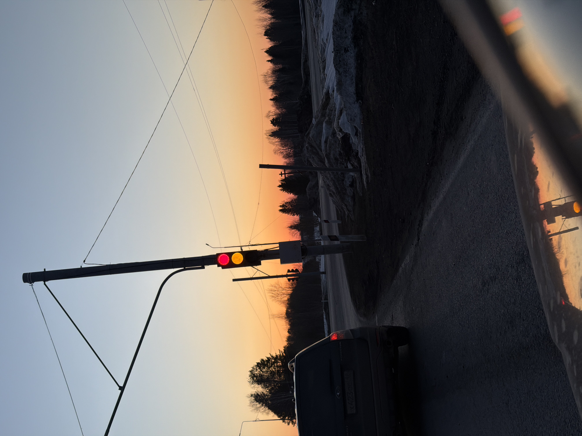 An image captured at an intersection in Shaberdino, Russia, depicts a twilight or dawn scene with a gradient sky transitioning from light blue at the zenith to orange-yellow near the horizon. In the foreground, a dark-colored Subaru Forester SUV, bearing a Russian license plate "X 423 OE 44," is stationary on an asphalt road, facing away from the viewer. The road appears damp or wet, with visible puddles and reflections. To the right of the SUV, a tall traffic light pole displays both red and yellow signals illuminated simultaneously. Overhead utility wires traverse the sky, and another streetlight is visible near the traffic light pole. On the right side of the road, significant piles of dirty snow and ice are present, interspersed with dark, bare ground. A dense line of mixed evergreen and deciduous trees forms the horizon, silhouetted against the sky. A wooden pole with a cross-like appendage is visible amongst the snow piles. The image perspective is from inside a vehicle, evidenced by the reflection of the sky and traffic light on a dashboard or hood in the lower frame.