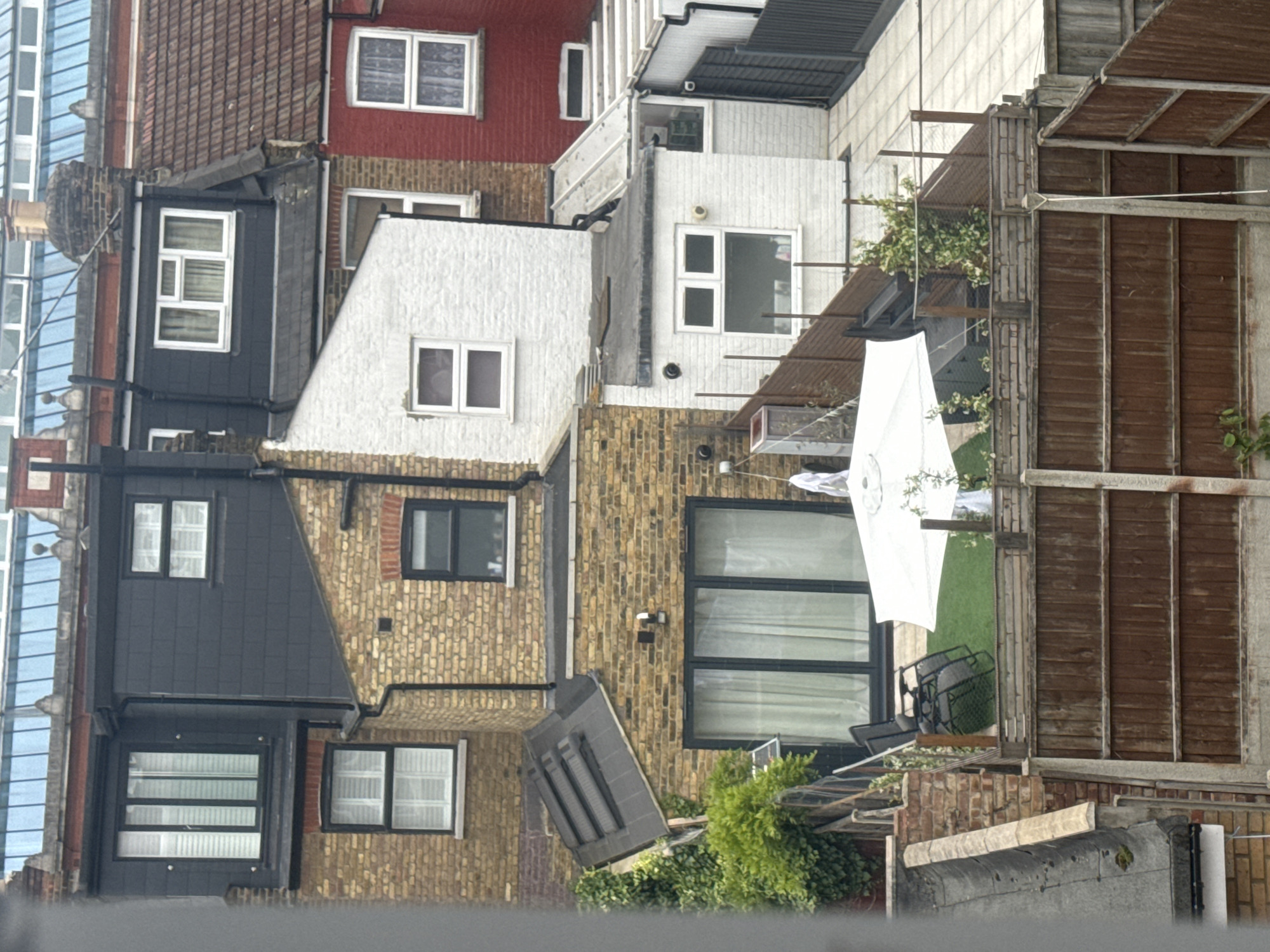 The image shows an aerial view of a backyard area between terraced houses. The houses are made of brick and painted white, black, and red, with pitched roofs. The backyard is divided into several sections by wooden fences. In the foreground, there is a wooden fence with concrete posts. Beyond that, there is a patio area with artificial grass, a white parasol, and some chairs. There are also several plants and shrubs in the garden. In the background, there are more terraced houses with windows and pitched roofs. The sky is overcast.
