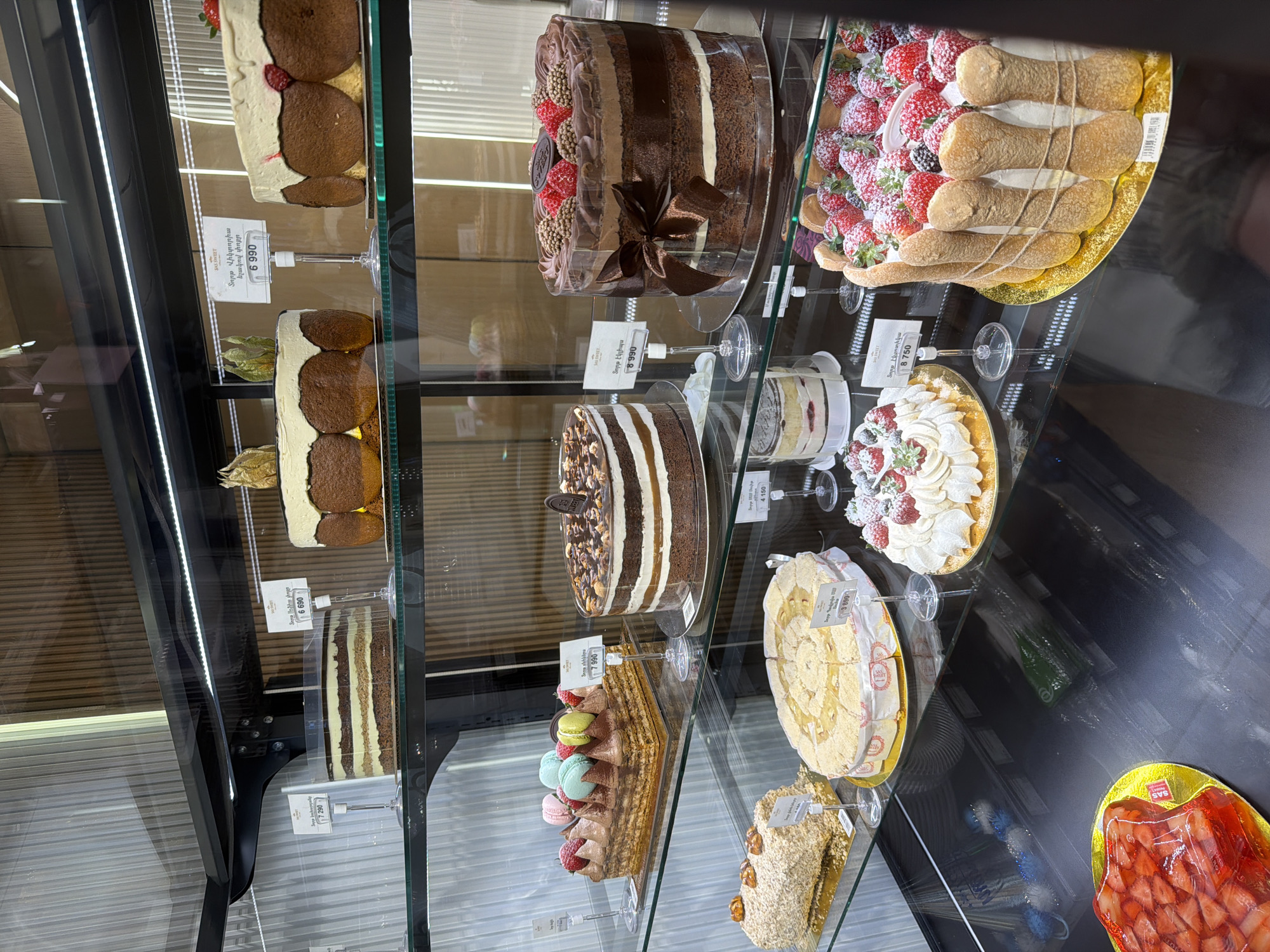 The image displays a brightly lit glass display case filled with an assortment of cakes and pastries, suggesting an indoor setting like a bakery or supermarket dessert section. The items are neatly arranged on multiple shelves, reflecting off the glass.
In the foreground, various multi-layered cakes are visible, including a dark chocolate cake with white cream layers and strawberries, and another cake with alternating dark and light layers. A large, round chocolate cake with a dark brown ribbon, decorated with red berries and chocolate sprinkles, is prominent. Next to it, a substantial dessert features ladyfingers surrounding a generous pile of strawberries dusted with powdered sugar. Other offerings include a round tart topped with white cream and berries, and several other multi-layered cakes, some topped with nuts, others with colorful macarons and strawberries. A brightly colored red dessert, possibly strawberry jelly, is partially visible in the bottom right corner.
No people are visible, and there's no discernible specific activity beyond the presentation of desserts for sale. The time of day and weather cannot be determined, as the scene is indoors with artificial lighting.
Several price tags are attached to the glass shelves. Many display the brand "SAS SWEET" and prices in Armenian Dram (AMD). Visible Armenian text identifies several cakes, such as "Տորթ Վիկտորիա ելակով փոքր" (Victoria cake with small strawberries) priced at 6,990 AMD, "Տորթ էկլիպս" (Eclipse cake) at 8,990 AMD, and "Տորթ Շոկոլադե Պրահա" (Chocolate Prague cake) at 8,750 AMD. Other prices include 7,290 AMD, 7,990 AMD, 4,150 AMD, and 3,890 AMD. The reflections in the glass obscure some details but generally show a clean, modern display environment.