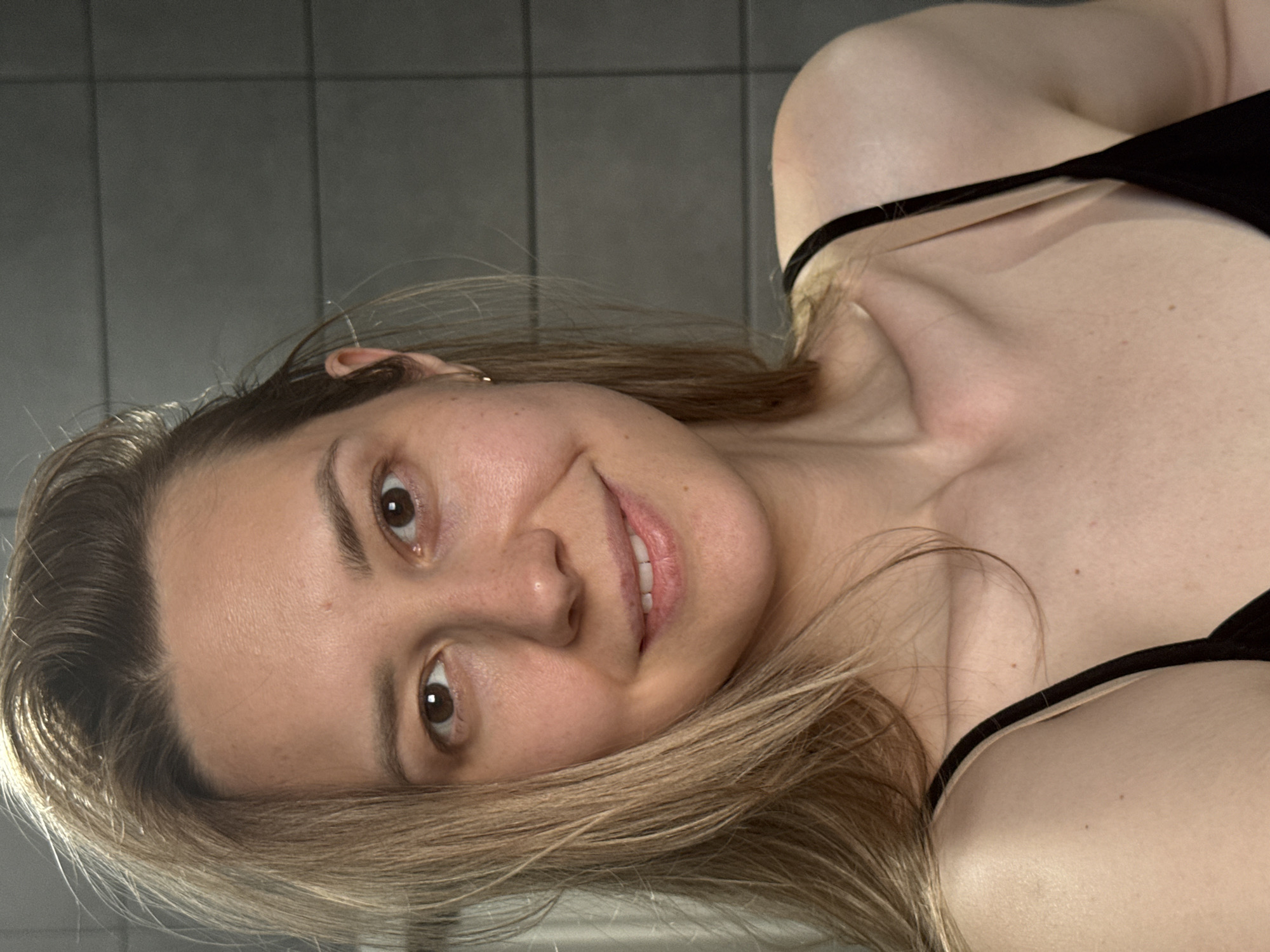 A selfie of a woman with brown hair and eyes, wearing a black tank top. The background shows light grey tiled walls. The location is not discernible as Chaikovsky, Russia from the provided information.
