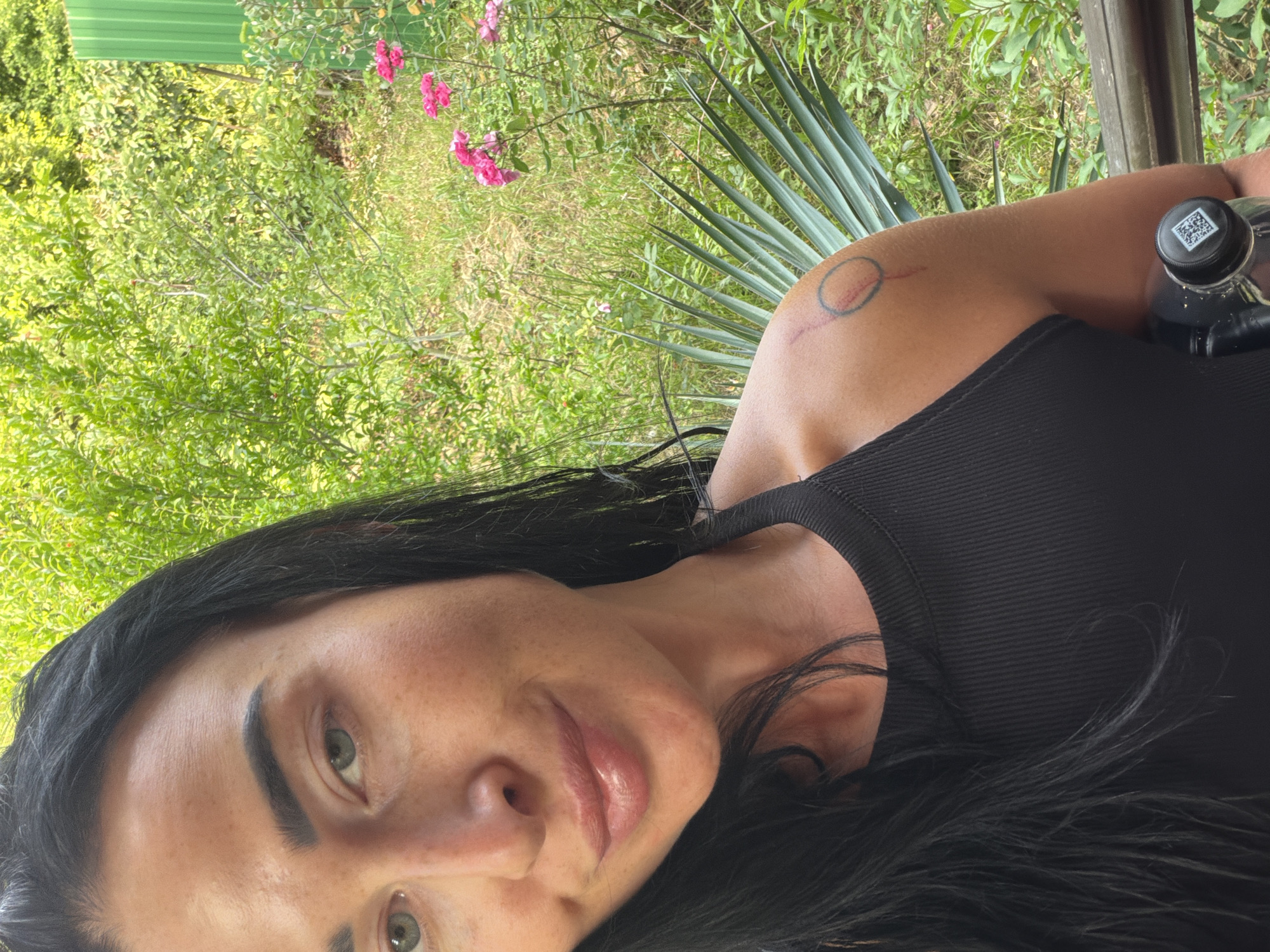 A selfie-style medium shot shows a woman with dark hair and light green eyes, wearing a black tank top. She has a tattoo on her right shoulder, appearing as a circle with a line extending from it. She is holding a black water bottle with a QR code on the cap. The background is lush with green foliage, including a palm frond, and pink flowers. The scene suggests an outdoor setting, potentially a garden or park. The image composition implies the woman is standing or sitting on a wooden structure, possibly a balcony or deck, overlooking the greenery. The lighting indicates it is daytime, with natural light illuminating the scene. The overall atmosphere is relaxed and casual. The location is identified as Sochi, Russia, though specific landmarks are not visible in the frame.
