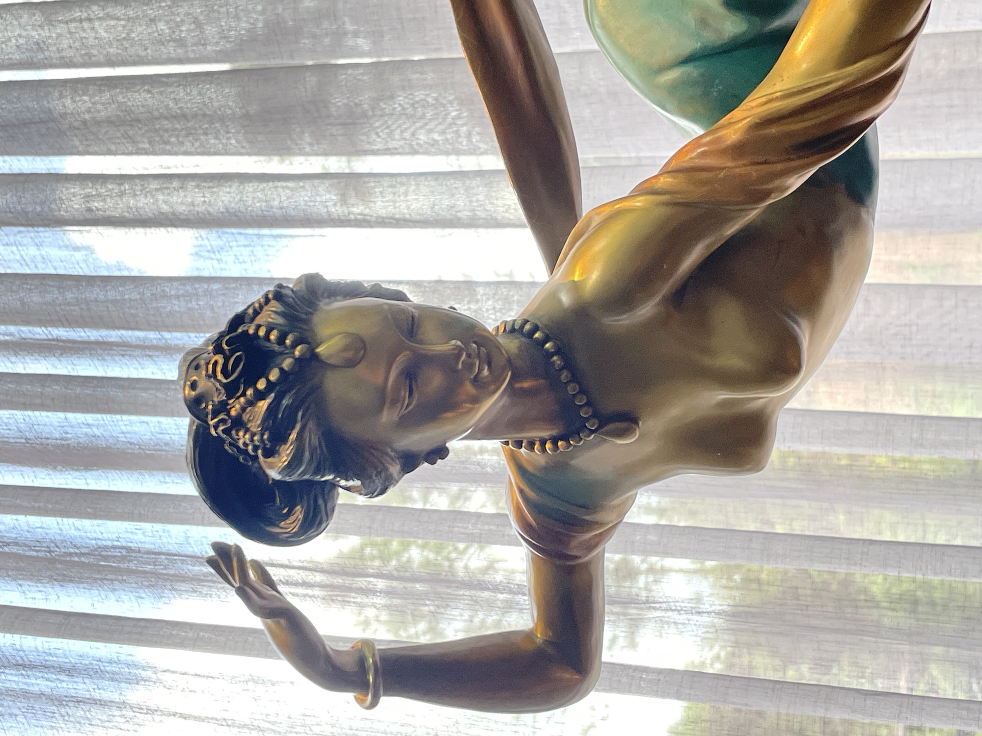 This is a close-up, vertically oriented photograph of a bronze sculpture of a woman, viewed from the chest up. The sculpture is ornate, with the woman depicted wearing a beaded necklace and an elaborate, dark-colored hairstyle adorned with gold embellishments. Her eyes are closed, and her head is tilted downwards, suggesting a state of contemplation or repose. Her right arm is bent, with her hand outstretched and fingers splayed, adorned with a golden bangle on her wrist. The sculpture's surface has a metallic sheen, with variations in color ranging from gold to a greenish patina, particularly on the body and a large, spherical object held or supported by the figure.
The background consists of translucent, horizontal blinds, behind which sunlight streams through, creating a soft, diffused light. The texture of the blinds is clearly visible, with horizontal folds and a fabric-like appearance. Through the blinds, faint hints of greenery and light suggest an outdoor scene, possibly trees or foliage, illuminated by daylight. The overall impression is one of peacefulness and artistry. No visible text is present in the image.