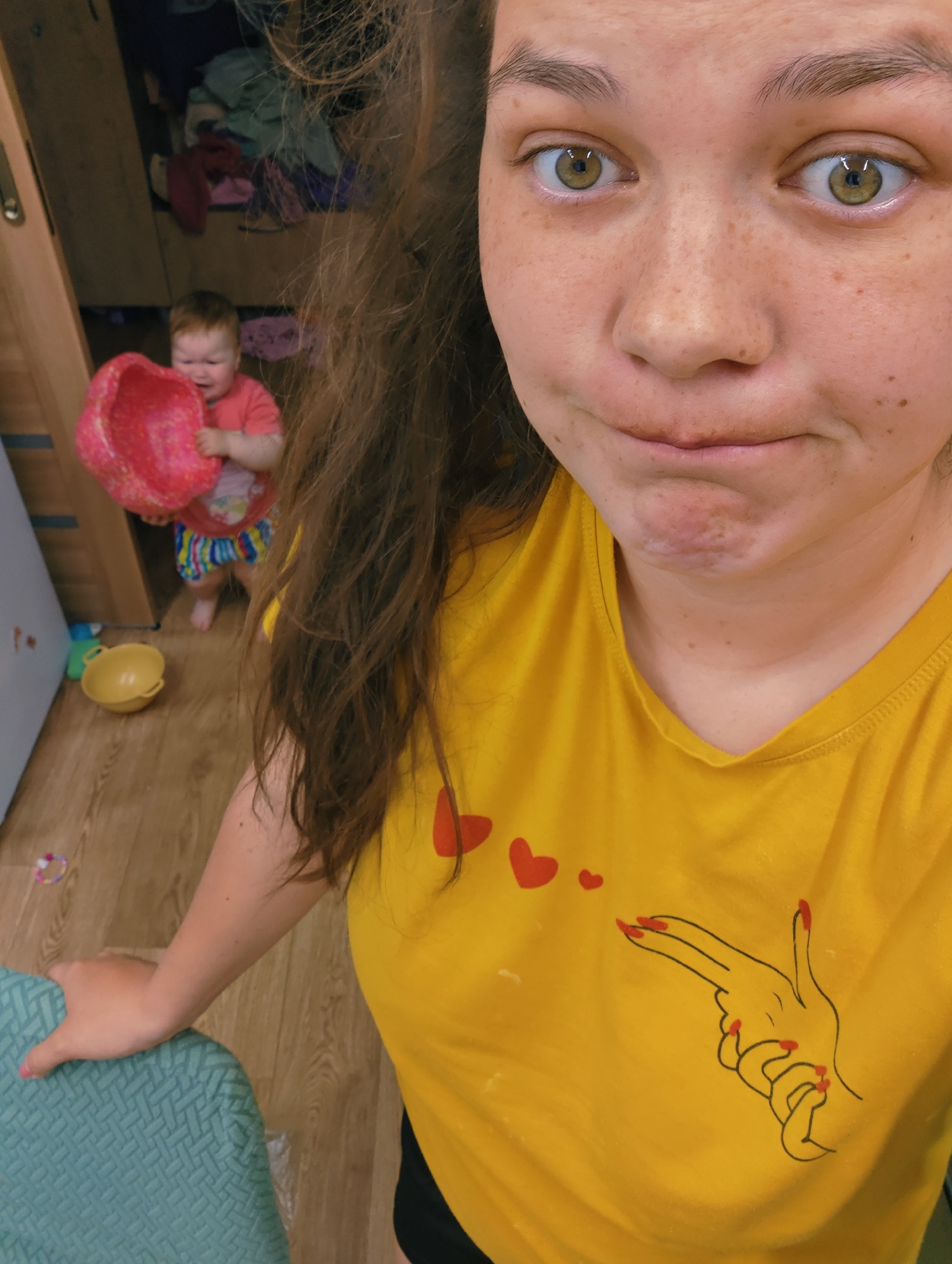 A woman is taking a selfie in what appears to be a home interior. She has light skin, freckles, and hazel eyes. Her hair is long and wavy, with some strands partially obscuring her face. She is wearing a yellow t-shirt with a hand-gun symbol and small red hearts printed on the front. Her expression is somewhat quizzical or amused.
In the background, slightly out of focus, a toddler is visible holding a pink plastic basket. The toddler is wearing a red shirt and multicolored striped shorts. A yellow plastic bowl is on the floor near the toddler. Behind the toddler is an open closet with clothing and other items visible inside.
The location is in Chaikovsky, Russia.
