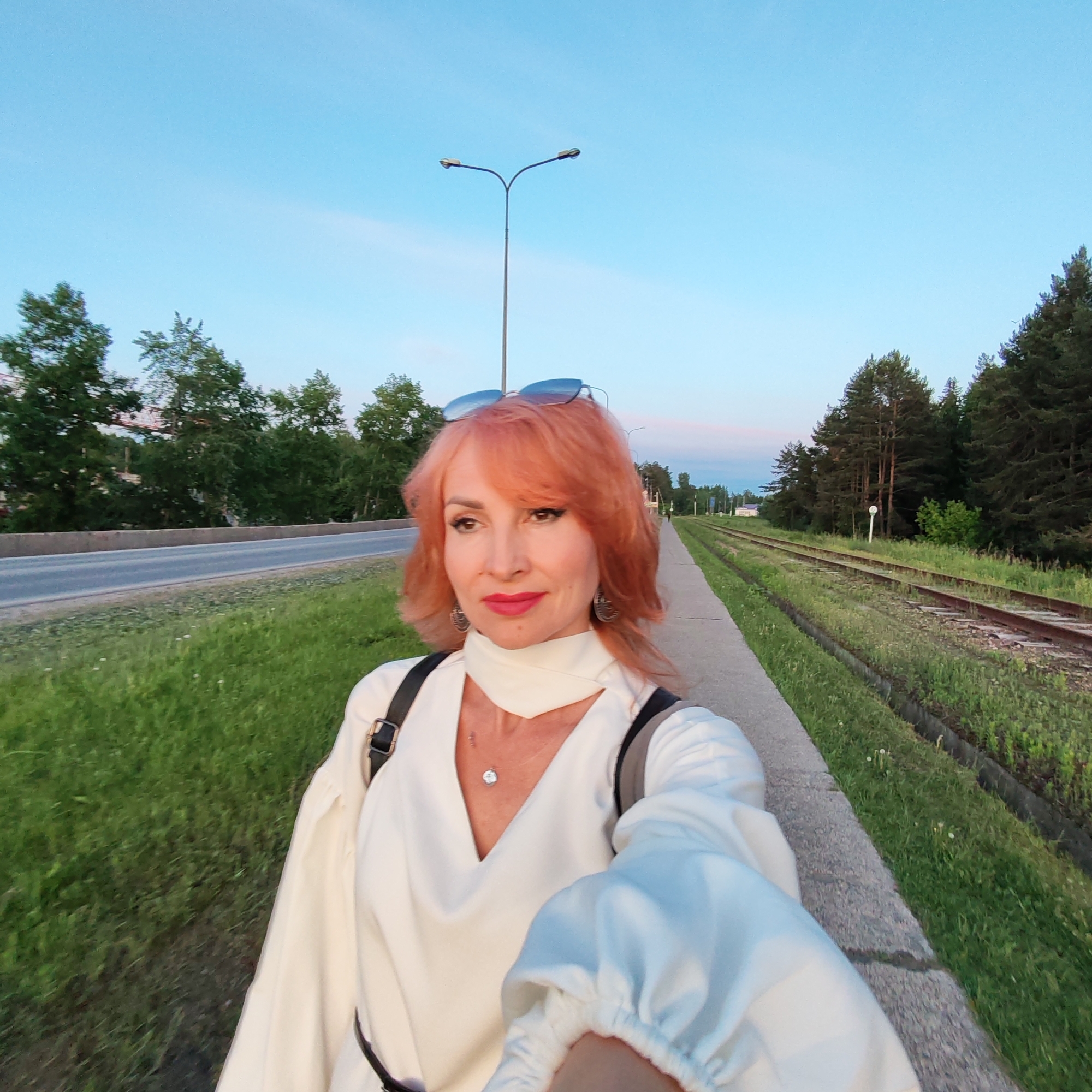 A woman with reddish-orange, shoulder-length hair and sunglasses perched on her head is captured in a selfie-style shot outdoors. She wears a white, long-sleeved top with a distinctive V-neckline and a high, draped neckband, accessorized with a silver pendant and ornate earrings. Her lips are adorned with red lipstick, and she has subtle eye makeup. She carries a dark-strapped item, possibly a backpack or bag, over her shoulders. Her expression is calm and composed as she looks slightly to her left.
The setting appears to be along a road or path in Chaikovsky, Russia. To the woman's left is an asphalt road, bordered by a concrete curb and green grass, with a line of trees visible in the distance. A tall street light pole stands prominently against the sky. To her right, adjacent to the sidewalk she stands on, are disused or infrequently used railroad tracks, overgrown with grass and weeds, flanked by a dense line of tall evergreen trees. The sky is a clear, light blue with soft hues of pink and orange on the horizon, indicating either early morning or late evening, commonly known as the "golden hour." The overall atmosphere is serene and natural, despite the proximity of infrastructure like the road and railway. No legible text is visible in the image.