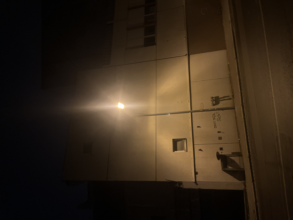 This nighttime photograph captures a stark urban building facade, likely located in Bamberg, Germany, illuminated by a single, prominent street light. The light, positioned towards the center-left, emits a strong, warm yellow-orange glow, creating a significant lens flare and brightly illuminating the light-colored, paneled surface of the building. The surrounding areas, particularly the sky to the left, are cast in deep shadow, emphasizing the artificial light source.
The building's facade is composed of large, rectangular panels, showing seams and subtle textures. Several small, dark, square-shaped openings or windows are visible across the illuminated wall. Above the main section, glimpses of what appear to be upper floors or more windows can be seen in the dim light. A thin pole extends horizontally from the light fixture, supported by another slender bar attached to the wall.
On the lower right section of the illuminated wall, there is dark graffiti. This includes a small, stylized, abstract drawing, possibly resembling a creature or symbol. Below this drawing, the German phrase "Zeit Für Sich!" is spray-painted in dark letters. This translates to "Time for oneself!" or "Time for yourself!". To the far right, a darker, vertical architectural element or an adjacent building wall frames the scene. The overall impression is one of a quiet, artificially lit streetscape after dark.