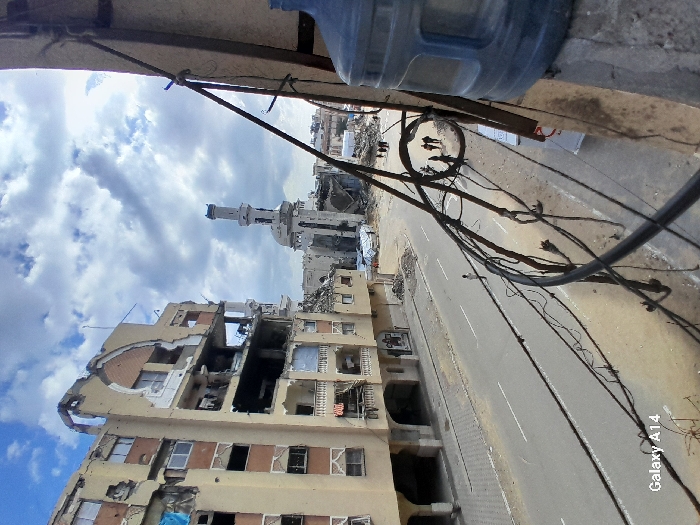 This image depicts a street scene in Khan Yunis, Palestinian Territory, characterized by extensive structural damage. In the immediate foreground, a partial view of a blue plastic water dispenser bottle is visible on the far right, resting on a concrete ledge. Numerous black electrical wires are strung irregularly across the right side of the frame, originating from an unseen attachment point on the right and extending towards the left and lower areas of the image.
The street, running from the mid-left to the upper right, is paved with asphalt and features faded white lane markings. On the left side of the street, a multi-story building, likely residential or commercial, shows severe structural damage, with large sections of its facade missing, exposing internal rooms and reinforced concrete. Windows are shattered or missing, and debris is visible on balconies and within the exposed interiors.
Further down the street, towards the center-right, an iconic mosque structure with a partially damaged white minaret is visible, surrounded by rubble and other heavily damaged or destroyed buildings. The mosque itself appears to have sustained significant structural compromise. Scattered piles of rubble and debris line the sides of the street.
Several individuals are present: one person walks alone in the middle of the paved road in the mid-ground, heading away from the viewer. Further down the street, near the mosque, a group of approximately five to seven individuals can be seen walking on the right side of the road, also moving away from the viewer. The sky above is partly cloudy, with a mix of blue sky and white and grey clouds. The overall environment suggests a scene of widespread urban destruction. The bottom left corner of the image contains a "Galaxy A14" watermark.