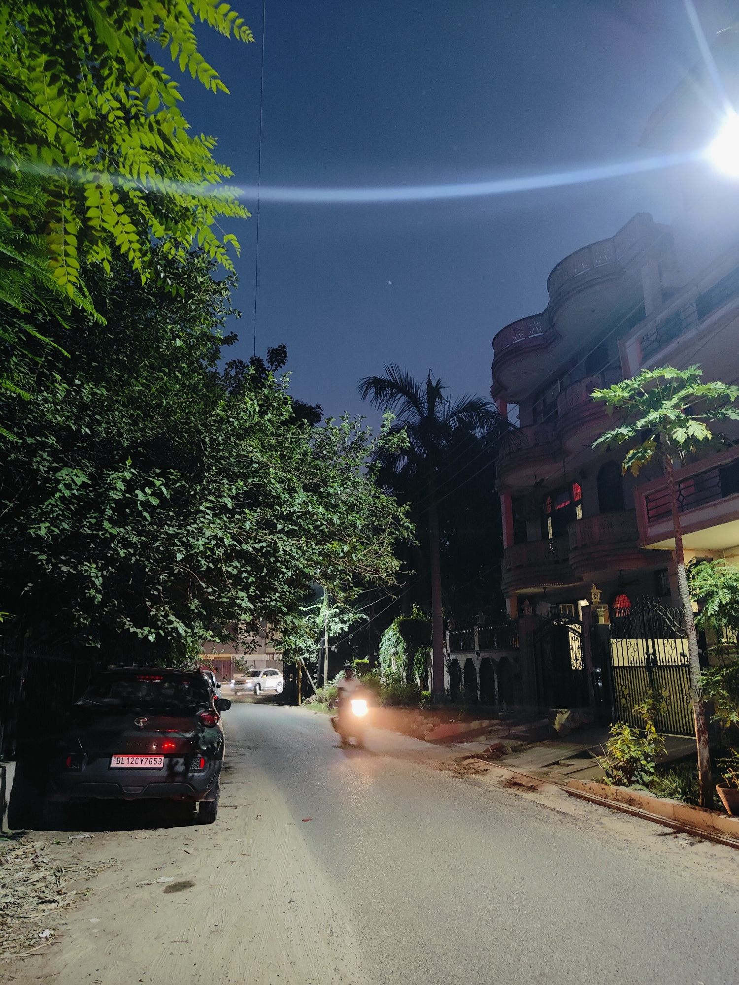 The image captures a street scene at night in Gurgaon, India, characterized by artificial lighting from streetlights and building interiors. In the foreground, a black car is parked on the left side of the road, with its license plate, "DL12CV7653," clearly visible. To the right of the car, the road curves slightly, illuminated by streetlights. A person on a motorcycle is visible moving away from the camera on the right side of the road, with their headlights on.
The environment includes lush green trees lining the left side of the street, their leaves illuminated by the artificial lights, creating a dappled effect on the ground. On the right side, a multi-story building with balconies and ornate architectural details stands prominently, its windows emitting a reddish glow. A fence and gate are visible in front of the building, with some greenery and potted plants. The sky is dark, suggesting it is late evening or night. The overall atmosphere is tranquil, with the primary activity being the passing motorcycle.