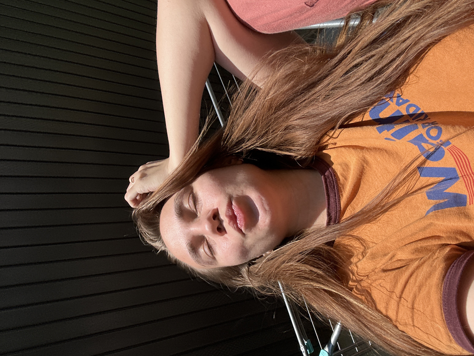 A woman with light skin and long, brown hair is lying down. She is wearing an orange t-shirt with blue and red lettering. Her eyes are closed and her lips are slightly parted. Her arm is raised above her head. The background includes a dark, horizontally-striped surface. Clothes drying rack is located near her body. The location is indiscernible.