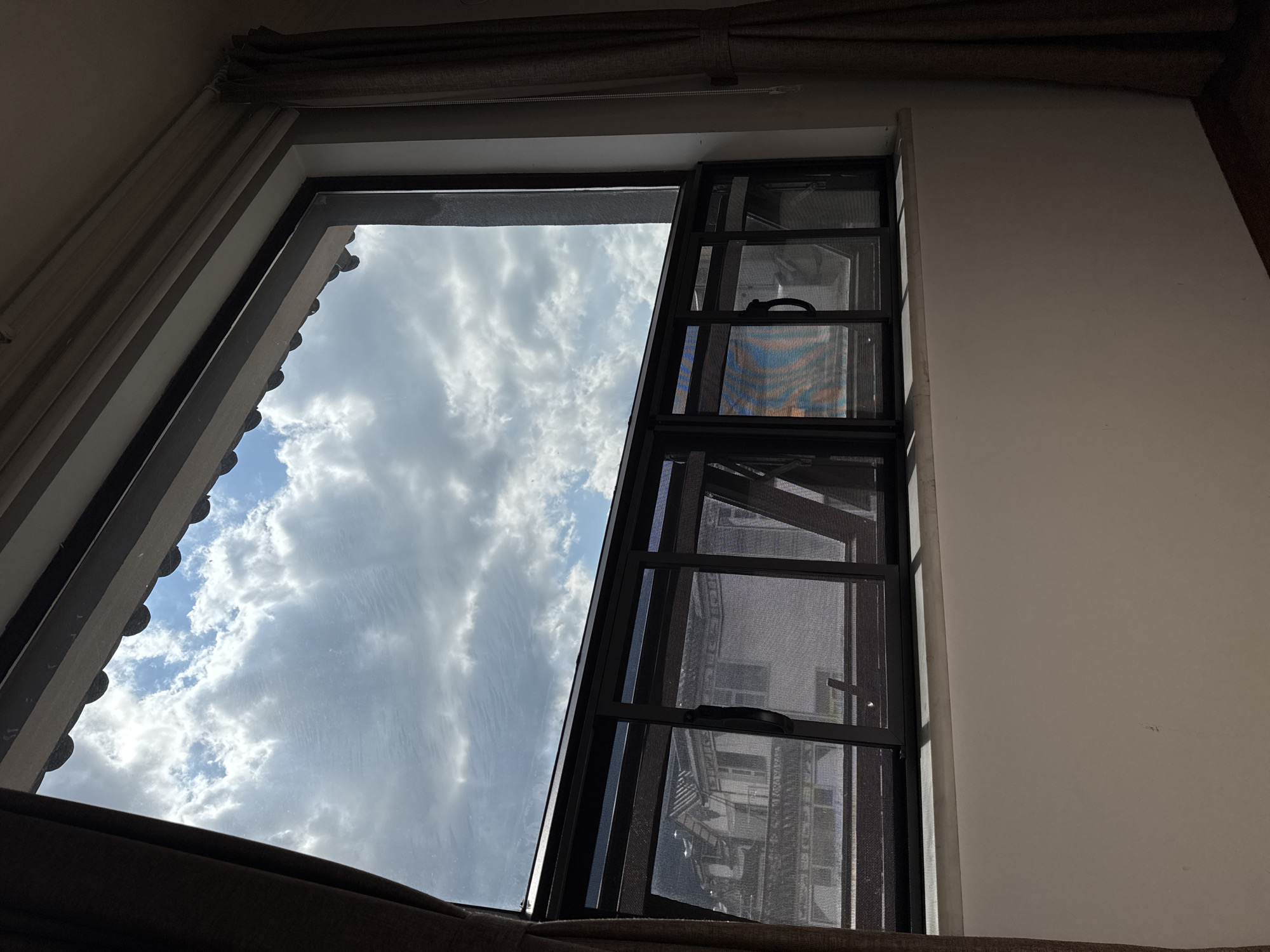 The image shows a view from a window, looking outwards towards a cloudy sky and buildings. The window has dark frames and a mesh screen. The sky is filled with white and gray clouds, with patches of blue sky visible. The buildings outside appear to be residential, with tiled roofs and balconies. The room inside the window is dimly lit, with curtains on either side. The scene is likely from a residential building in Dali City, China, given the architectural style of the buildings and the context provided. There are no people or animals visible in the image.