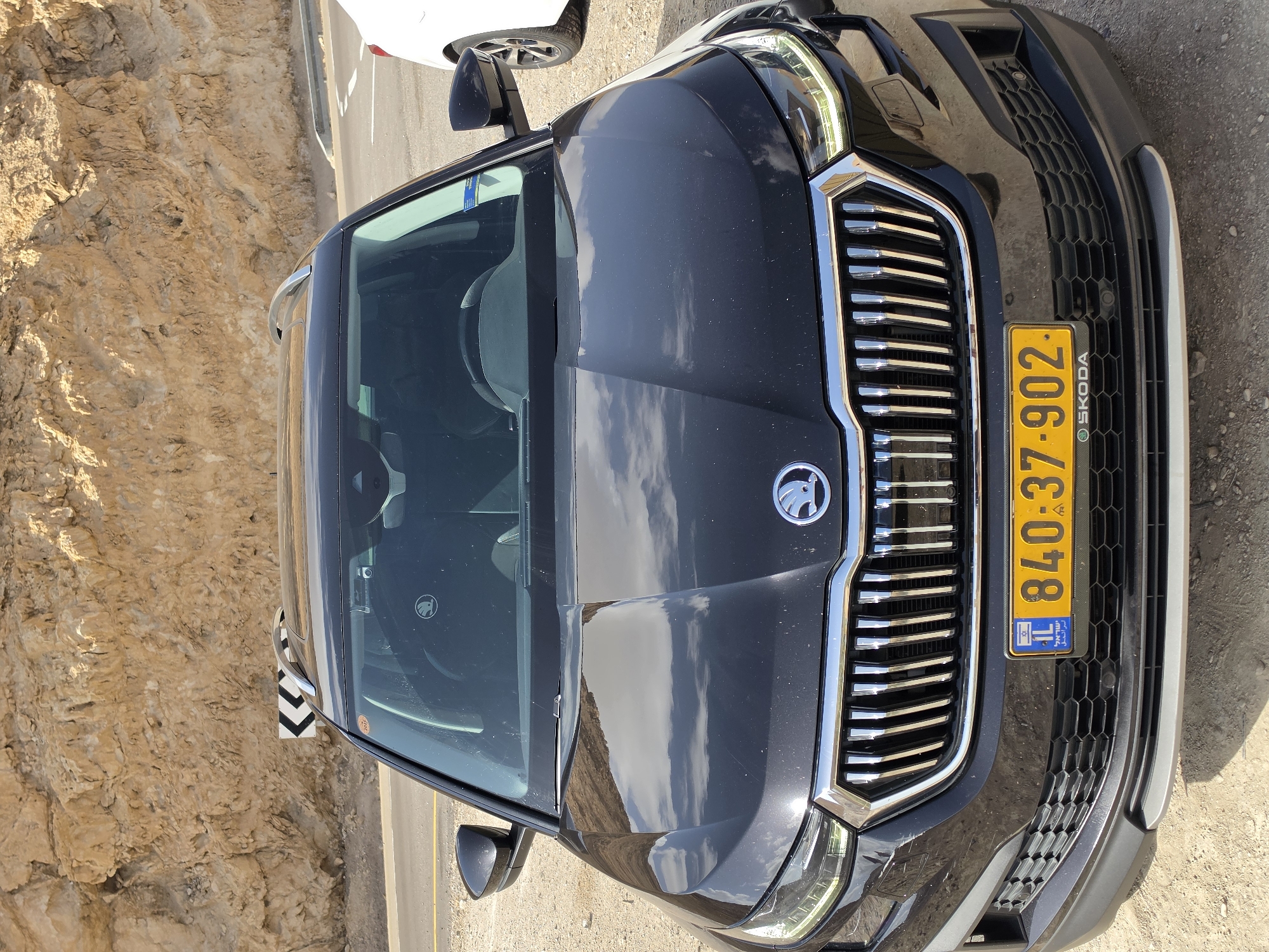 A dark gray Škoda car is parked on an unpaved surface next to a road with a rocky cliff visible in the background. The front of the car is in view, showing the Škoda emblem on the hood and a yellow Israeli license plate reading "840-37-902." The environment suggests a location within the Tamar Regional Council, Israel, given the license plate and arid landscape. No people are visible in the frame.