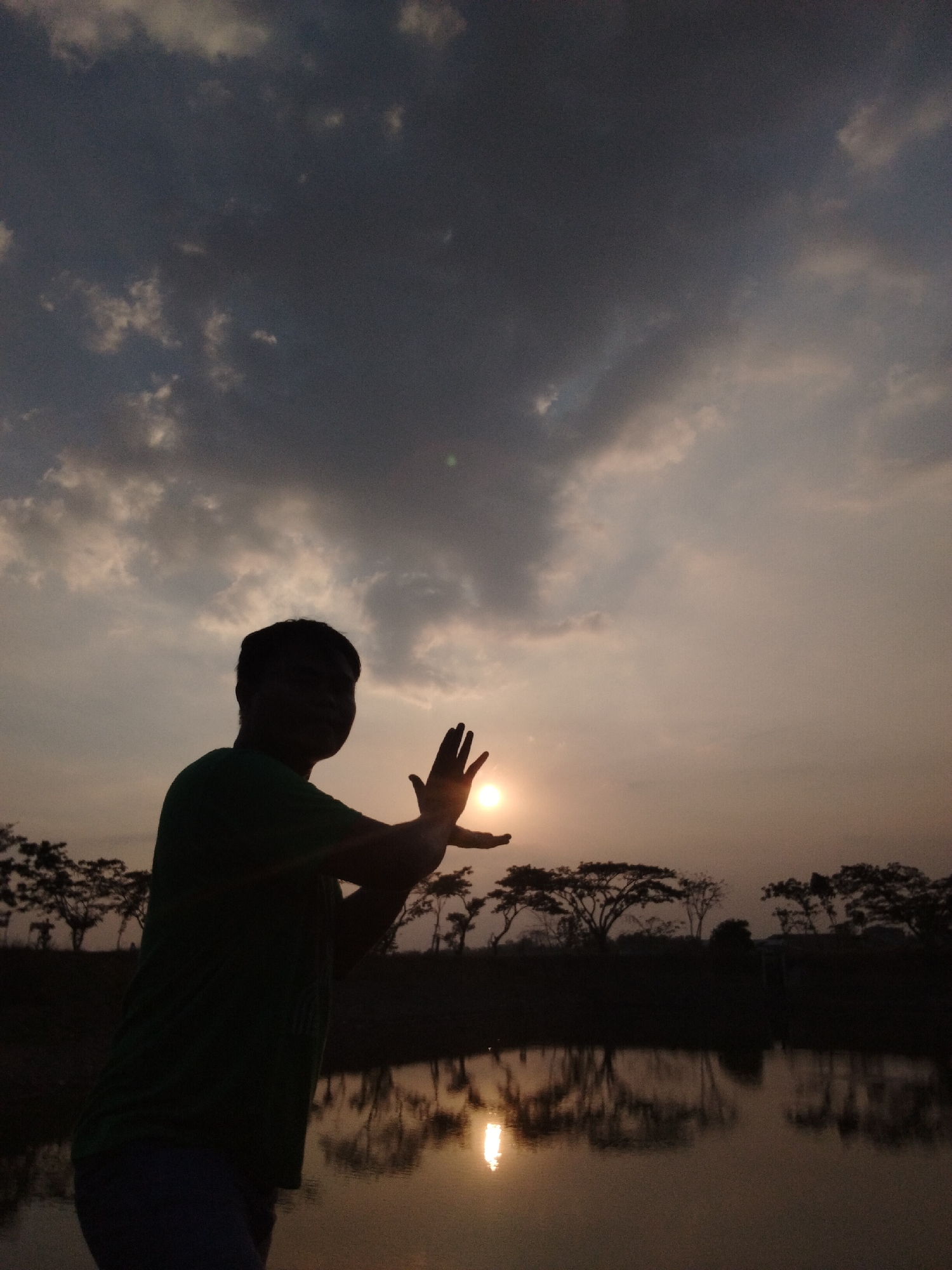 A silhouette of a person stands in the foreground against a dusky sky, with their hands held up as if framing the setting sun. The person is wearing a dark green t-shirt and dark pants. The sun, a bright orb, is visible just above the horizon, partially obscured by the person's hands. In the background, a line of trees forms a dark silhouette against the sky, and their reflections are visible in a calm body of water in the midground. The water's surface is smooth, creating a mirror image of the trees and the sky. The overall lighting suggests it is dusk or dawn.