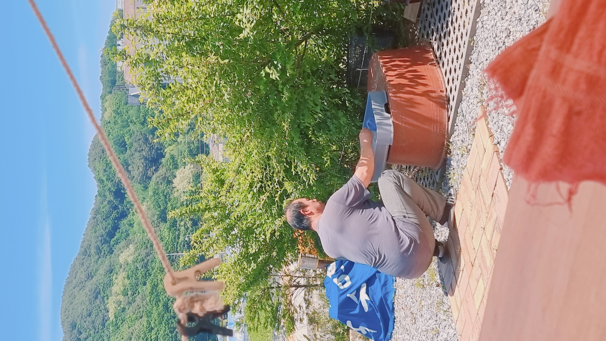 The image shows a man crouching next to a large, rust-colored container outdoors in Dunnae-myeon, South Korea. The man, with a receding hairline, wears a gray t-shirt and gray pants. He appears to be placing a gray tray-like object into the container. A blue banner with white lettering lies on the ground near him.
The scene is on a paved area of red bricks, surrounded by white gravel. In the background is lush greenery, possibly a hillside covered with trees, beneath a clear blue sky. A clothesline with clothespins is visible in the foreground. A portion of a red object is on the right edge.