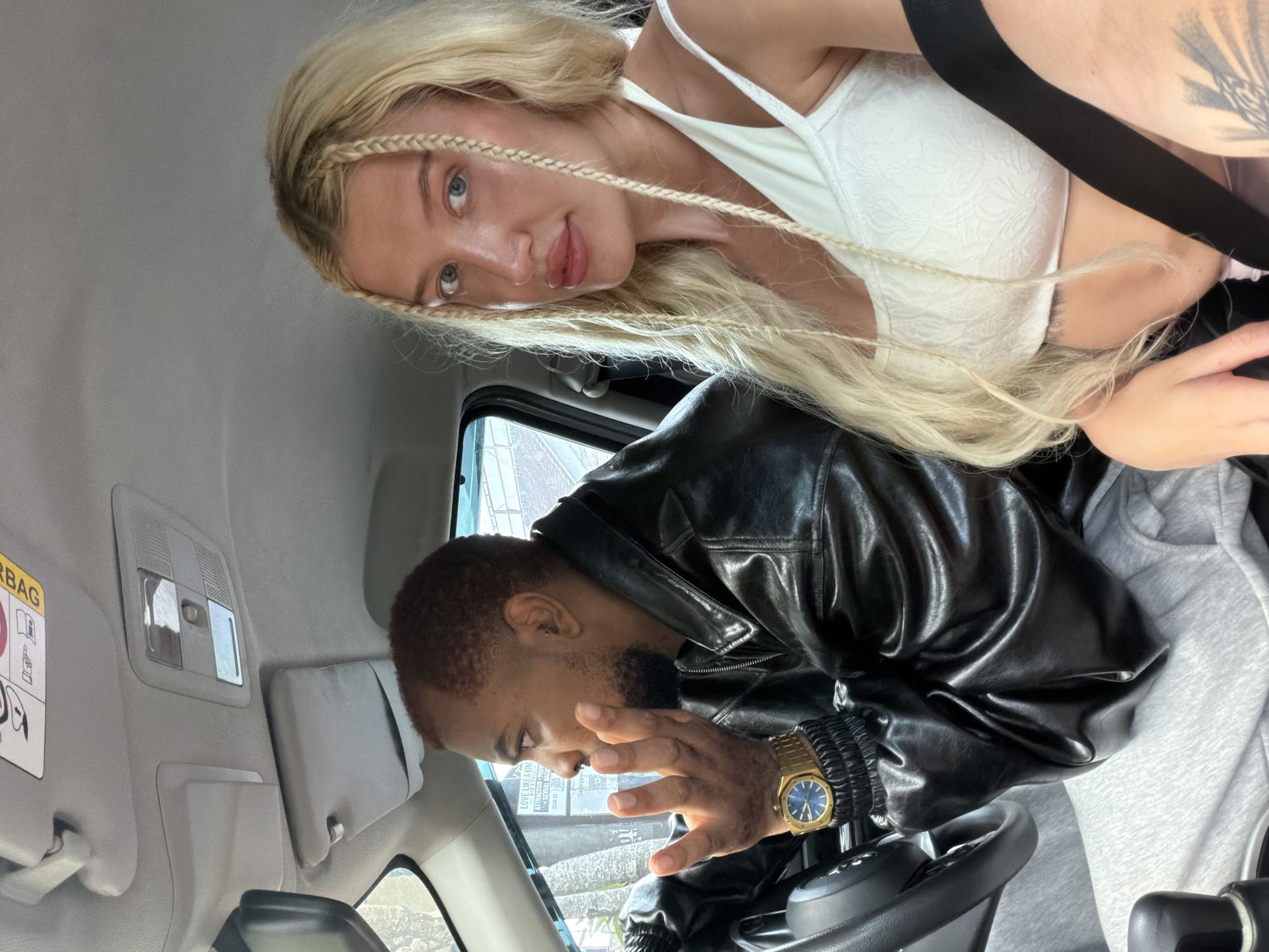 The image shows two individuals in the interior of a car, likely in the city of Mengwi, Indonesia. The person in the driver's seat is a man with reddish-brown hair and a beard, wearing a black leather jacket and a gold watch with a blue face. He is looking to his left with his right hand raised, appearing to be speaking or gesturing. The woman in the passenger seat has blonde hair styled in braids and is wearing a white top. She is looking at the camera. The car's interior includes the dashboard, a sun visor with a warning sticker, and part of the steering wheel. The background visible through the car windows suggests an outdoor urban or suburban environment.