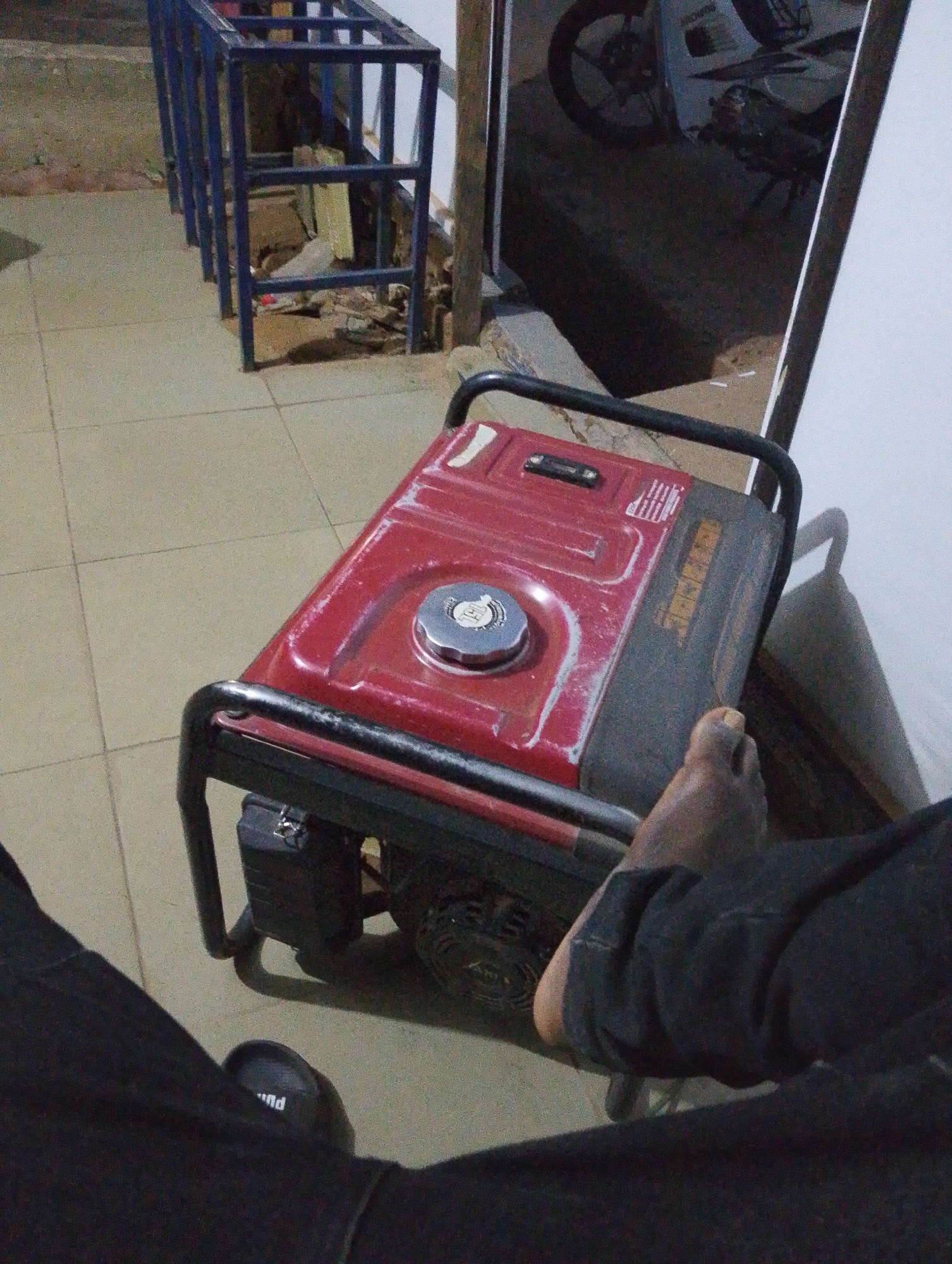In Sabon Tasha, Nigeria, a red generator sits on a tiled floor next to a blue metal frame structure. The generator has a black frame and a silver cap on its top. A person's foot, wearing dark clothing, is visible near the generator. In the background, a motorcycle is parked, and a white wall is present. The scene appears to be outdoors, possibly in a yard or open area.