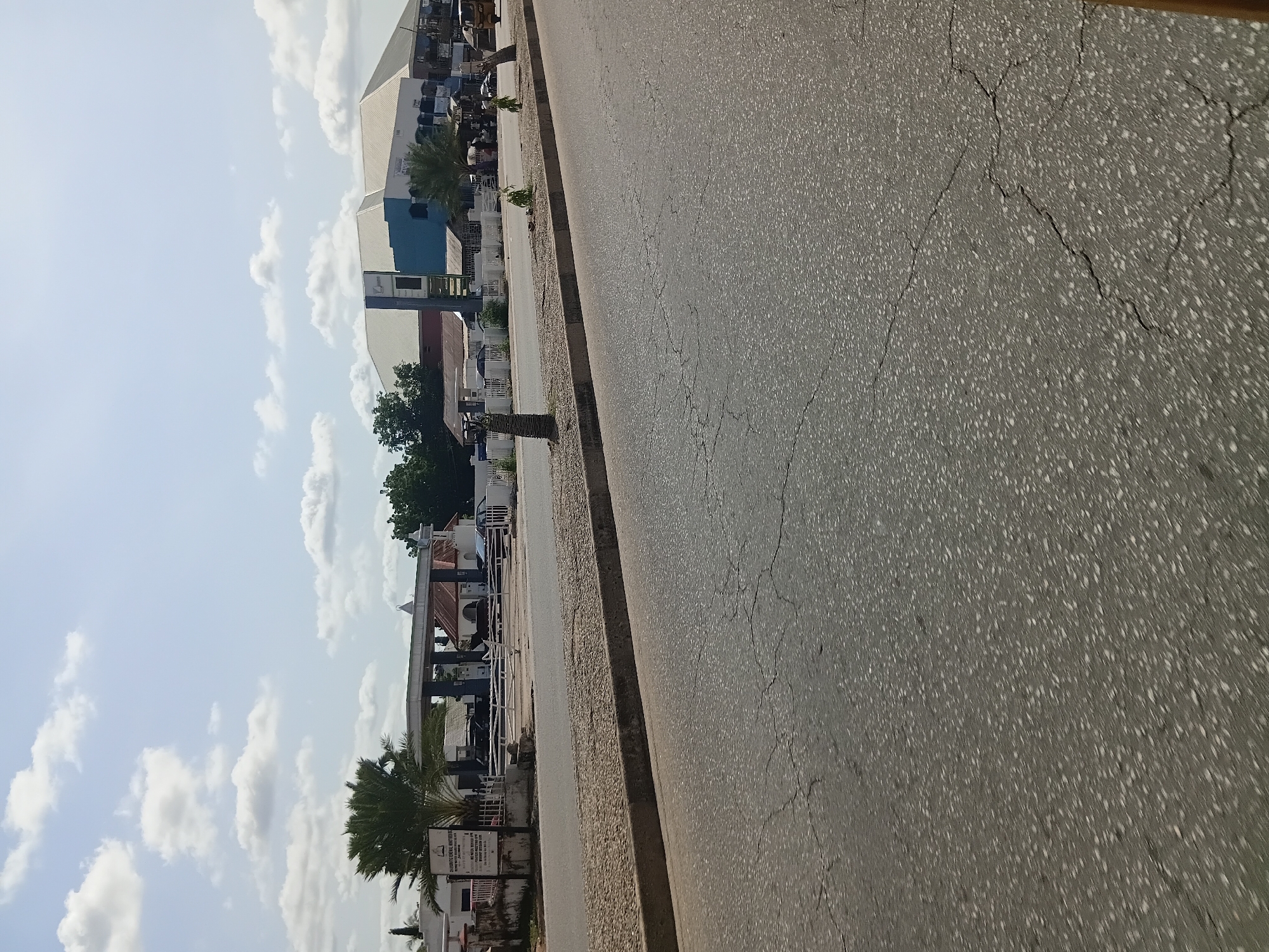 The image shows a street scene in Jalingo, Nigeria. In the foreground, a cracked asphalt road occupies most of the frame. A concrete curb separates the road from a gravelly median strip. Beyond the median, a white metal fence encloses a property with a gas station and several buildings. The gas station has a blue and white canopy over pumps, with cars parked nearby. To the right of the gas station, a large, modern building with a white and blue facade is visible. Palm trees are scattered throughout the scene, and the sky is partly cloudy. The scene appears to be a commercial area along a main road.