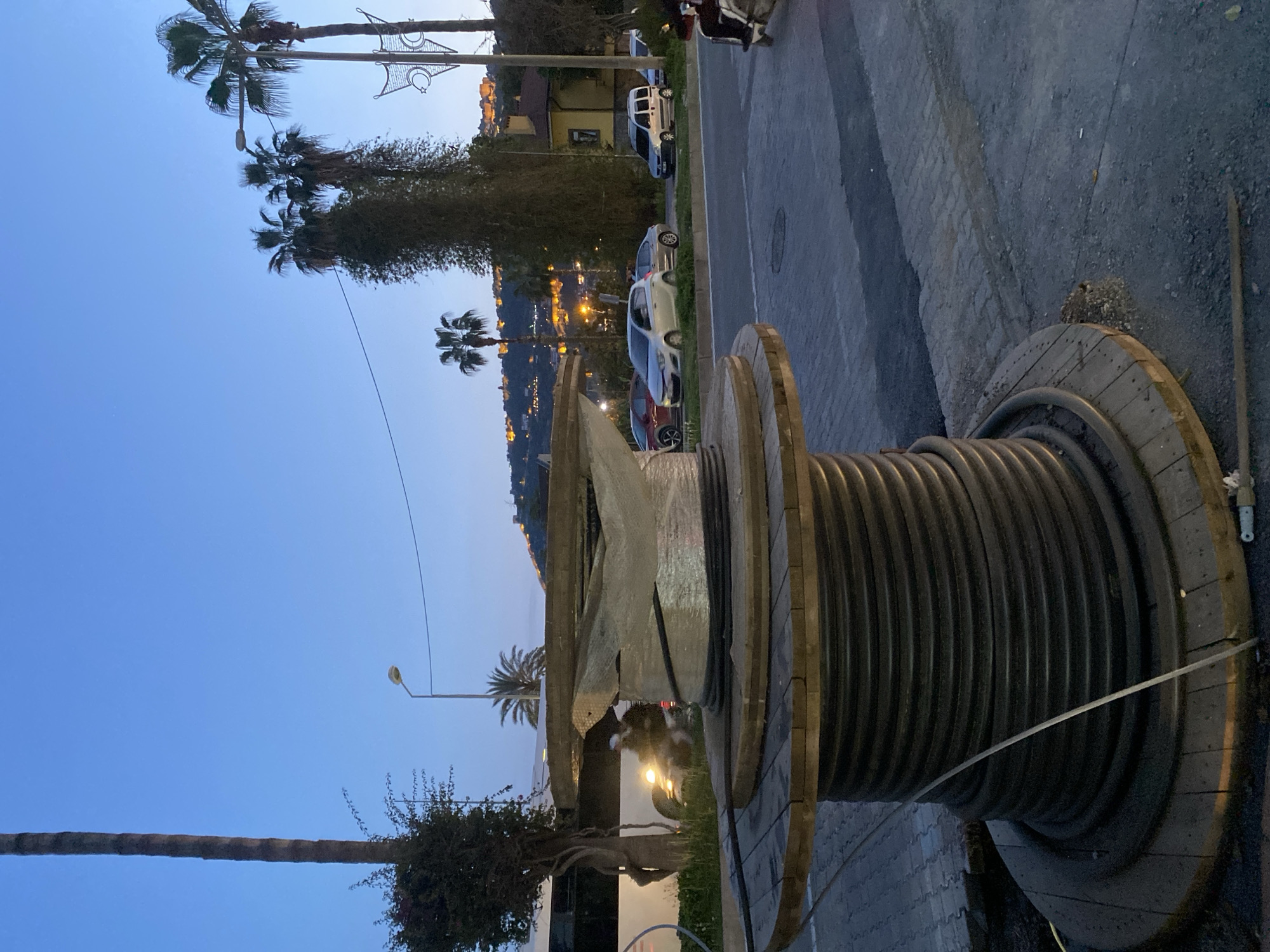 The image captures a street scene at dusk or early evening in Alanya, Turkey. The sky, visible in the upper left, is a clear, deep blue, fading slightly to a lighter hue towards the horizon.
In the foreground, dominating the right side of the frame, is a large, wooden cable spool. A thick, black cable is wound around its core, and a section appears to be unwound, indicating ongoing or recent work. The spool rests on a paved surface, likely a sidewalk or roadside.
To the left of the spool and extending into the midground, a street is visible, with several cars parked along its side. These include a white SUV, a grey hatchback, and a red car, suggesting a residential or commercial area. Interspersed among the parked vehicles and along the roadside are various trees and bushes, including several tall palm trees that dot the landscape and reach towards the sky. One decorative street light fixture, shaped like a crescent moon and star, is visible in the upper right, a strong cultural indicator of Turkey.
In the background, hills or mountains rise, covered with numerous buildings whose lights are illuminated, creating a warm glow against the twilight. This suggests a vibrant town or city built on elevated terrain. Another street light is visible against the sky in the mid-left, casting a soft glow.
No people are visible in the scene, and there is no discernible text. The overall atmosphere is tranquil, characteristic of an evening in a Mediterranean-style town.