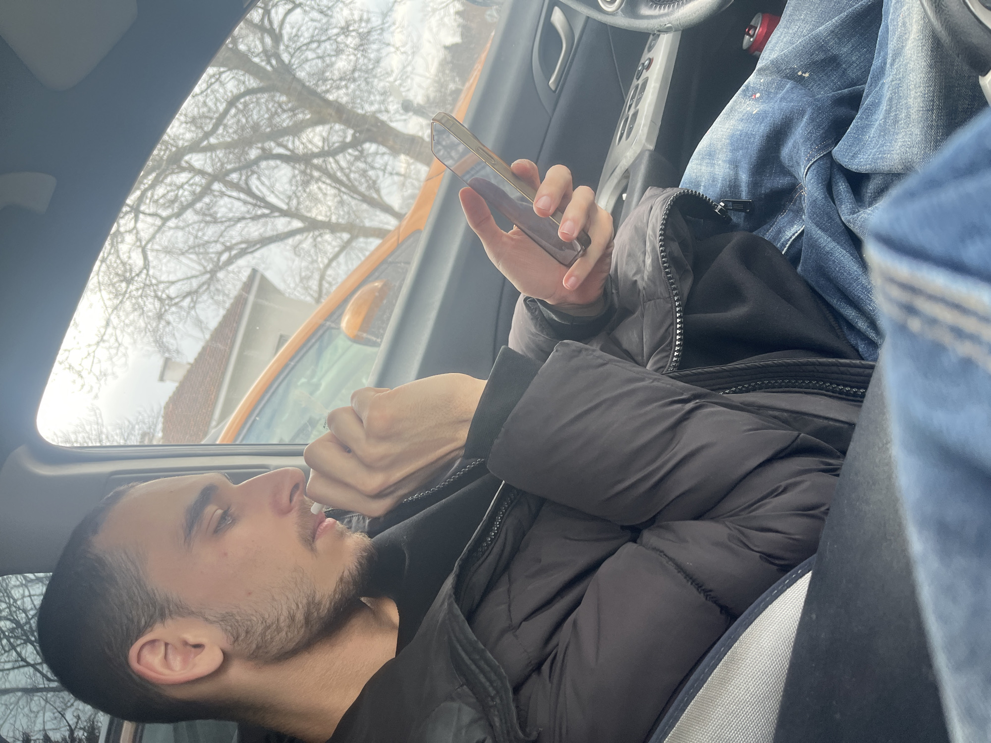 A young man with a trimmed beard is reclining in the passenger seat of a car. He is wearing a black puffer jacket and blue jeans. He is holding a smartphone in his right hand, appearing to be focused on the screen. Outside the car window, a leafless tree is visible against a cloudy sky. Part of an orange car is visible, also. The location is Varna, Bulgaria.